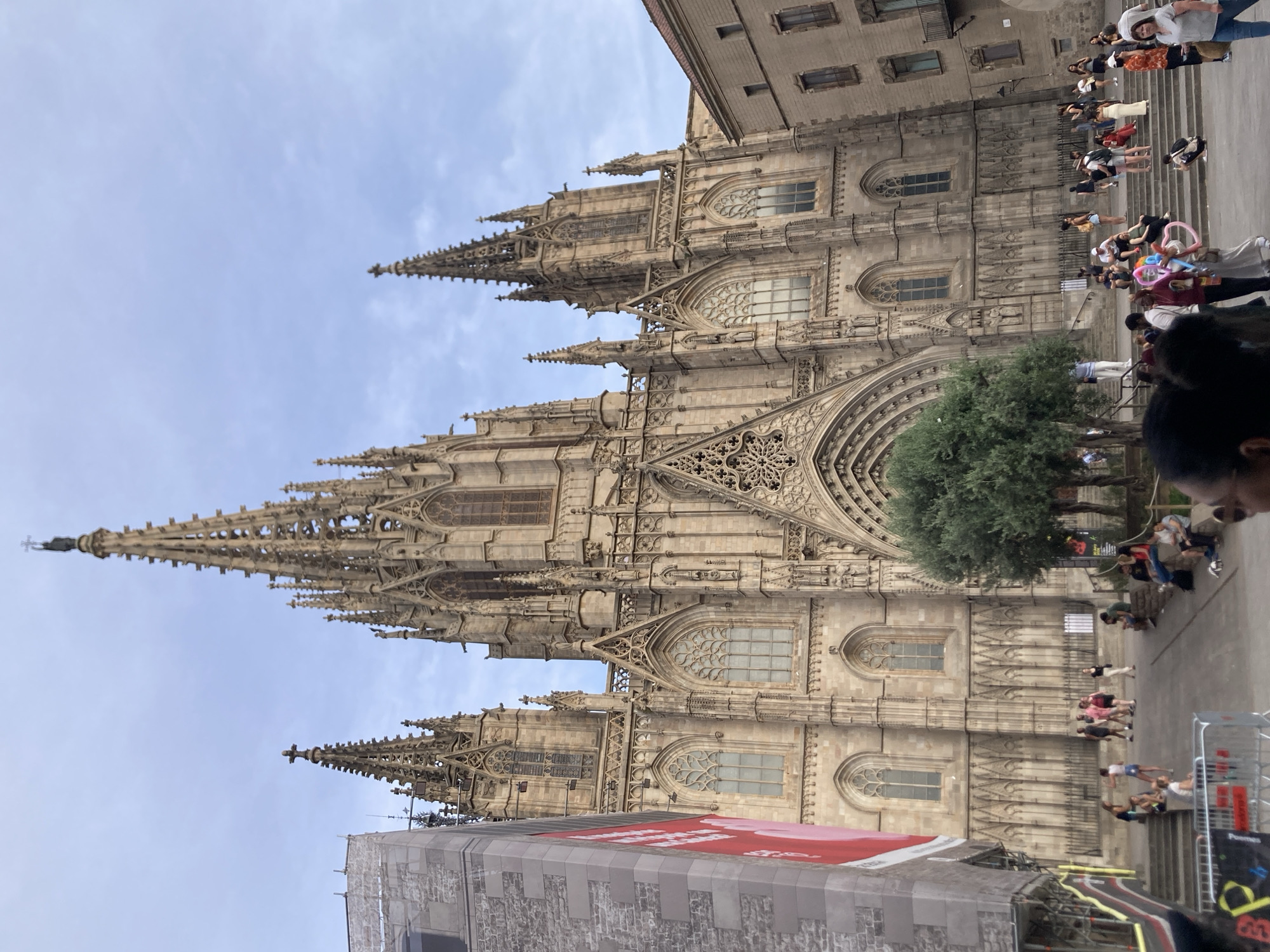 The image depicts a wide-angle, ground-level view of the Barcelona Cathedral in Barcelona, Spain. The cathedral's facade is a dominant feature, characterized by its Gothic architecture, intricate stonework, pointed arches, and soaring spires. A large, well-maintained tree is situated in front of the main entrance, adding a touch of greenery to the scene.
In the foreground and middle ground, a crowd of people is gathered. They are casually dressed and appear to be tourists and locals enjoying the public space. Some individuals are walking, while others are sitting on steps or the ground, observing the cathedral or interacting with each other. The presence of a red banner on a construction-like structure to the left suggests ongoing urban development or events.
The sky above is partly cloudy, indicating an outdoor setting during daylight hours. The overall atmosphere appears relaxed and communal, with the grand architectural structure serving as a backdrop to human activity. The scene captures a moment of everyday life in a historically significant urban environment.