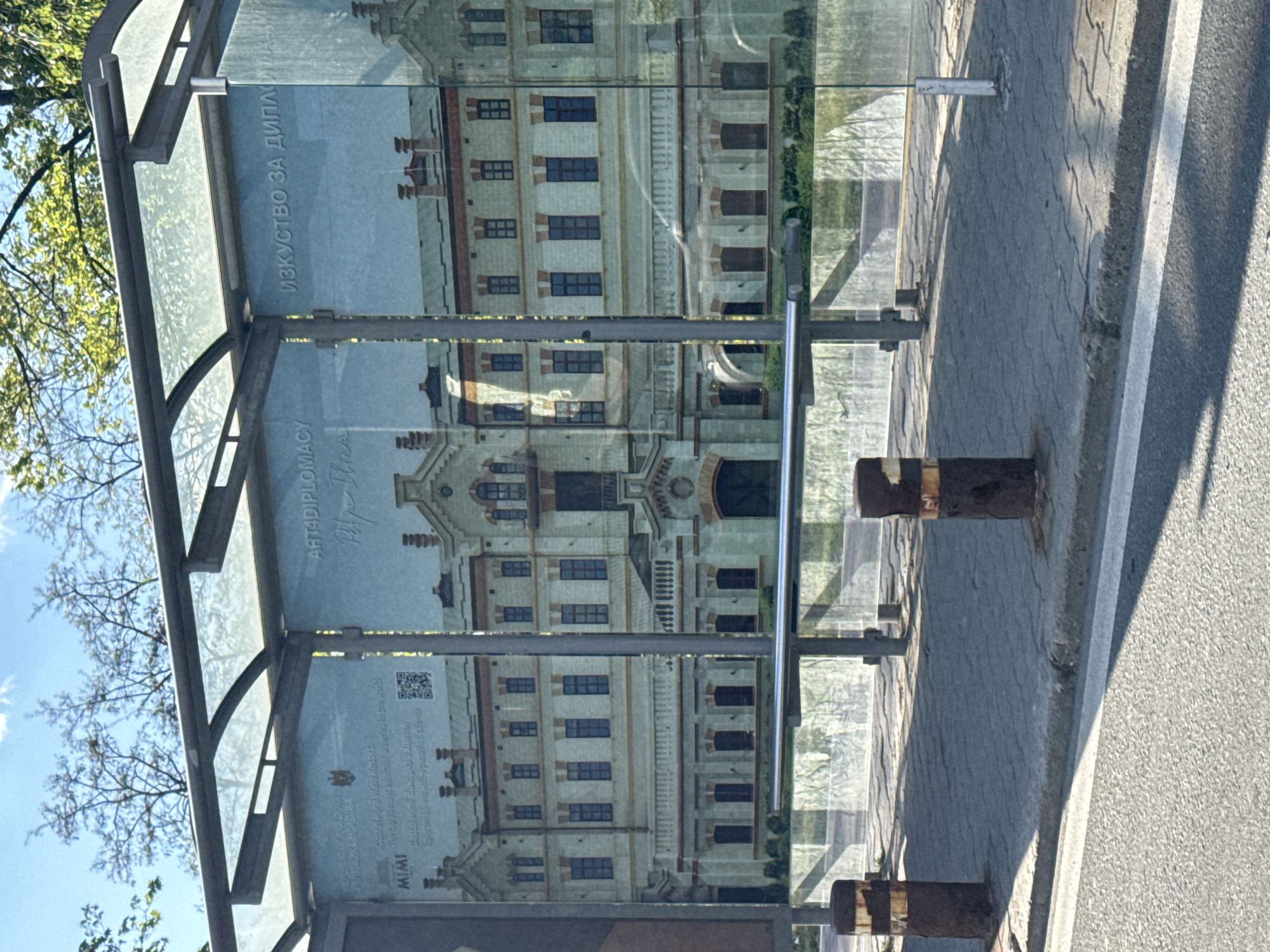 A bus stop in Sofia, Bulgaria, displays an advertisement for "ART4DIPLOMACY" featuring a large, vertically-oriented image of a white building with multiple windows and architectural details. The advertisement is partially reflected in the glass of the bus stop shelter. The environment includes trees, a sidewalk with paving stones, and a street. Rust-colored metal bollards are positioned near the bus stop.