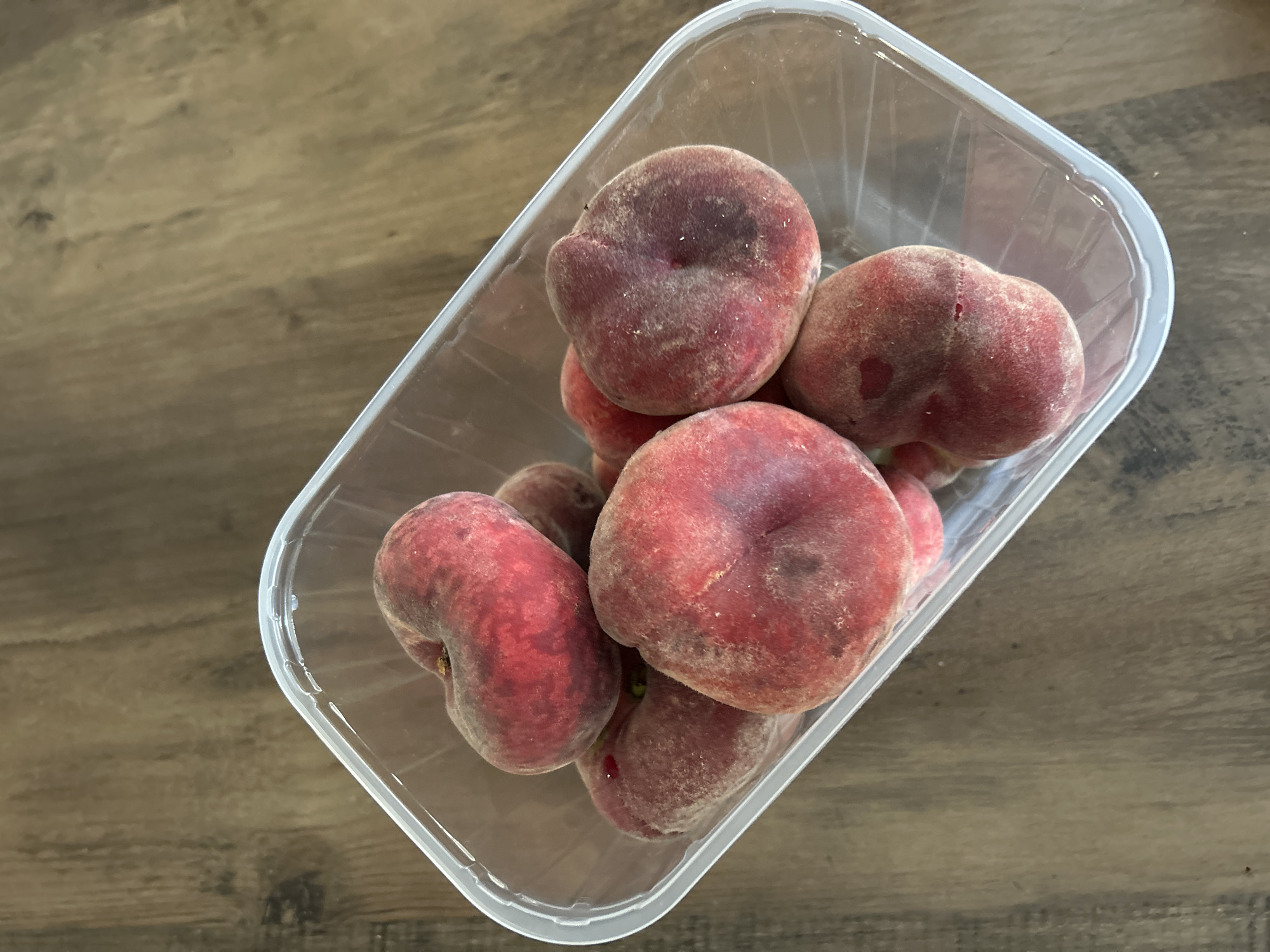A clear plastic container filled with several flat peaches sits on a wooden surface. The peaches are a mix of red and pink hues, with a fuzzy texture. The container has a clear lid that is partially ajar. The wooden surface appears to be a table or countertop with a visible grain pattern. The lighting suggests an indoor environment, likely a kitchen or pantry. The scene is static, with no apparent action or people present. The overall impression is one of fresh produce, possibly prepared for consumption or storage. The image was taken from a slightly elevated angle, looking down into the container.