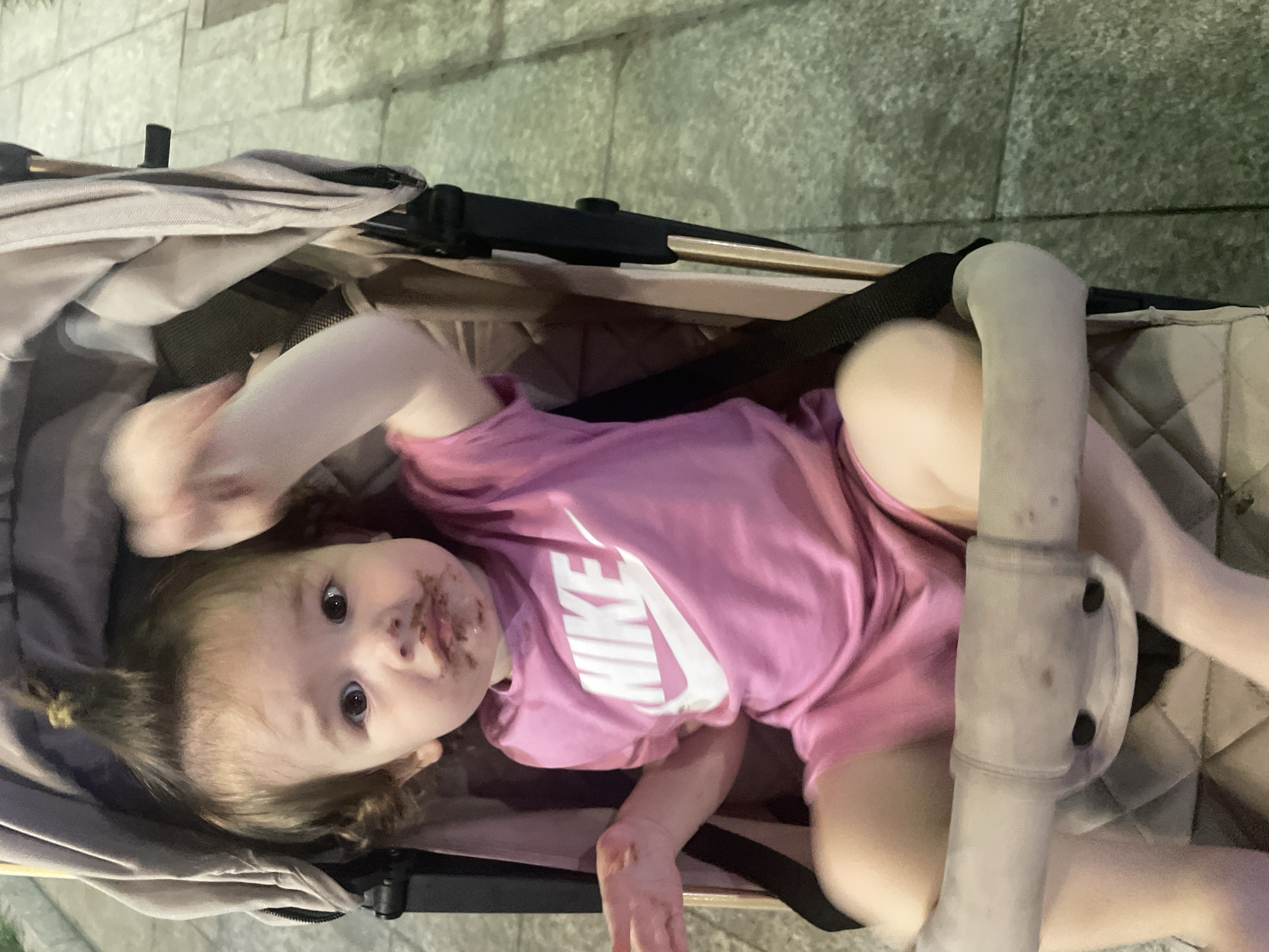 A toddler is sitting in a stroller. The toddler has chocolate smudged around their mouth and on their shirt. The toddler is wearing a pink t-shirt with a white Nike logo. The stroller is beige and black. The background is blurred and appears to be an outdoor setting with paved ground.
