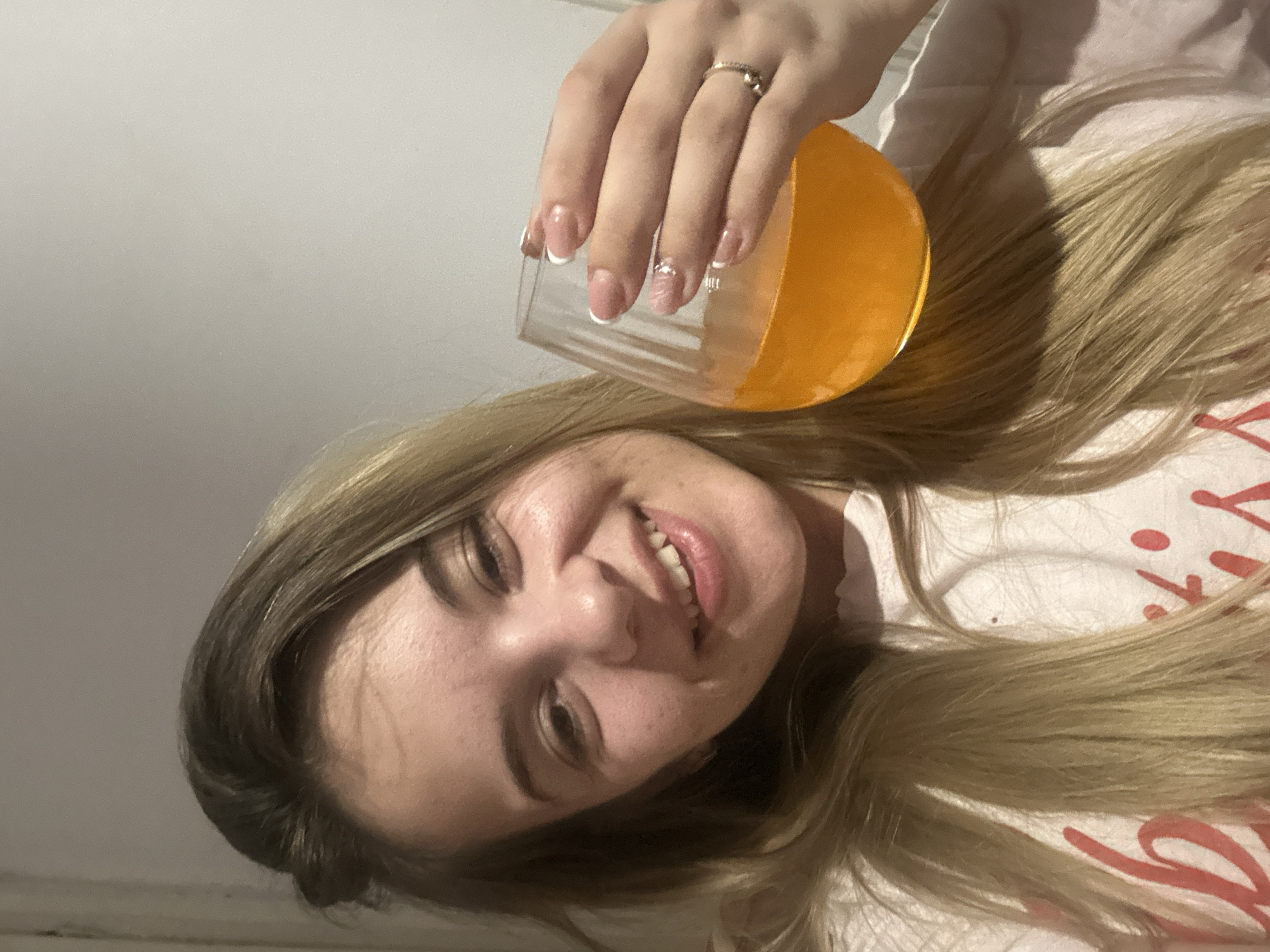 A fair-skinned woman with light brown hair and light-colored lipstick is holding a clear glass filled with an orange liquid. The glass is slightly tilted, and the woman's hand, adorned with a ring, is grasping it. She is smiling. She is wearing a light-colored shirt. The background is a plain wall and part of a white surface possibly the bedsheet. No information indicates the location is in Plovdiv, Bulgaria.