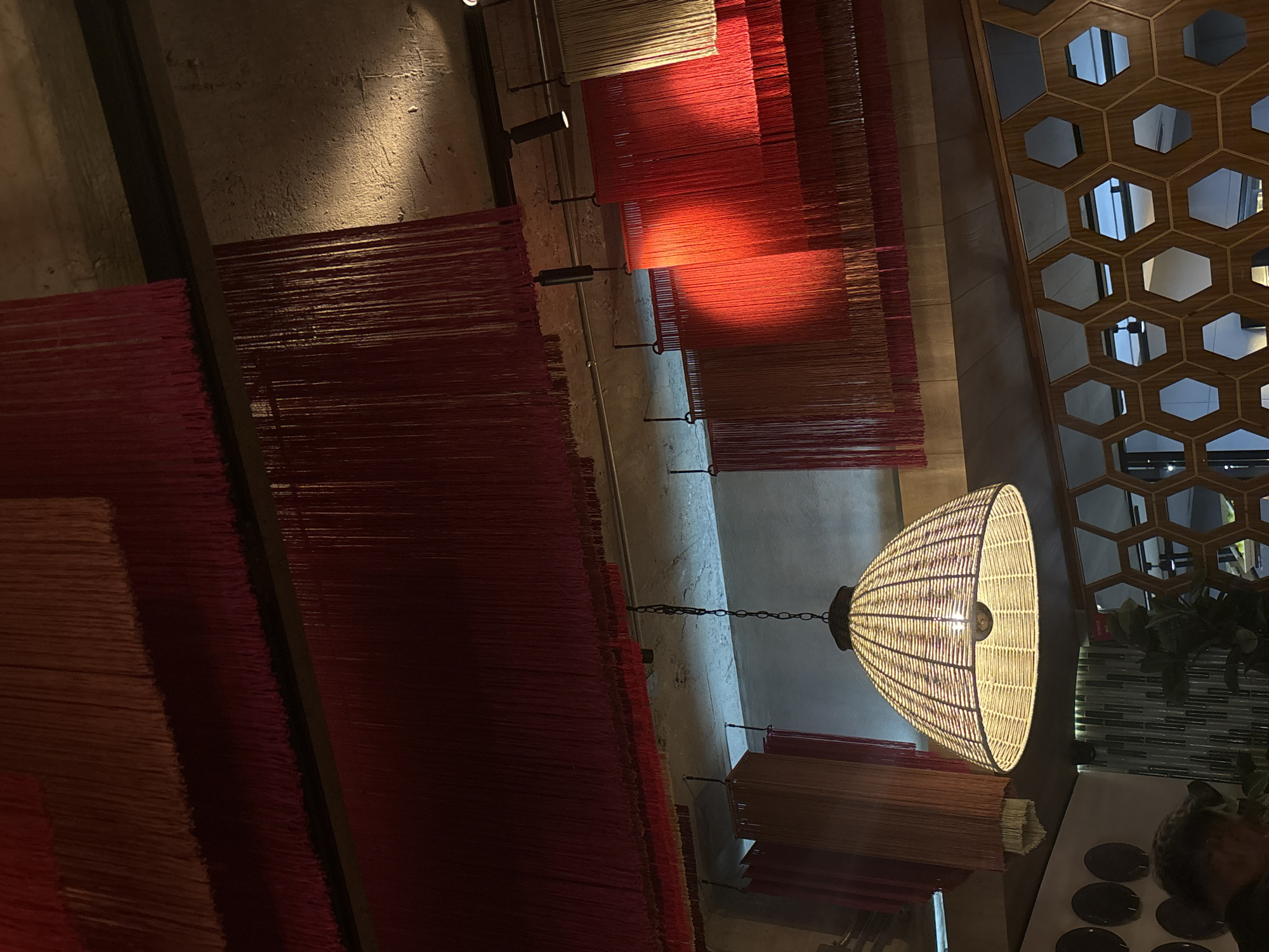The interior of a restaurant in Mexico City, Mexico, is captured in an eye-level shot. The ceiling is adorned with an installation of horizontal panels of vertically hanging thin strips of fabric in various shades of red, orange, and beige. Track lighting fixtures are visible. A large, cylindrical wicker pendant lamp is suspended in the center of the frame. To the right is a vertical, honeycomb-patterned screen with glimpses of the outside. A table setting with dishes and glassware is visible on the bottom right, with a person partially in frame.