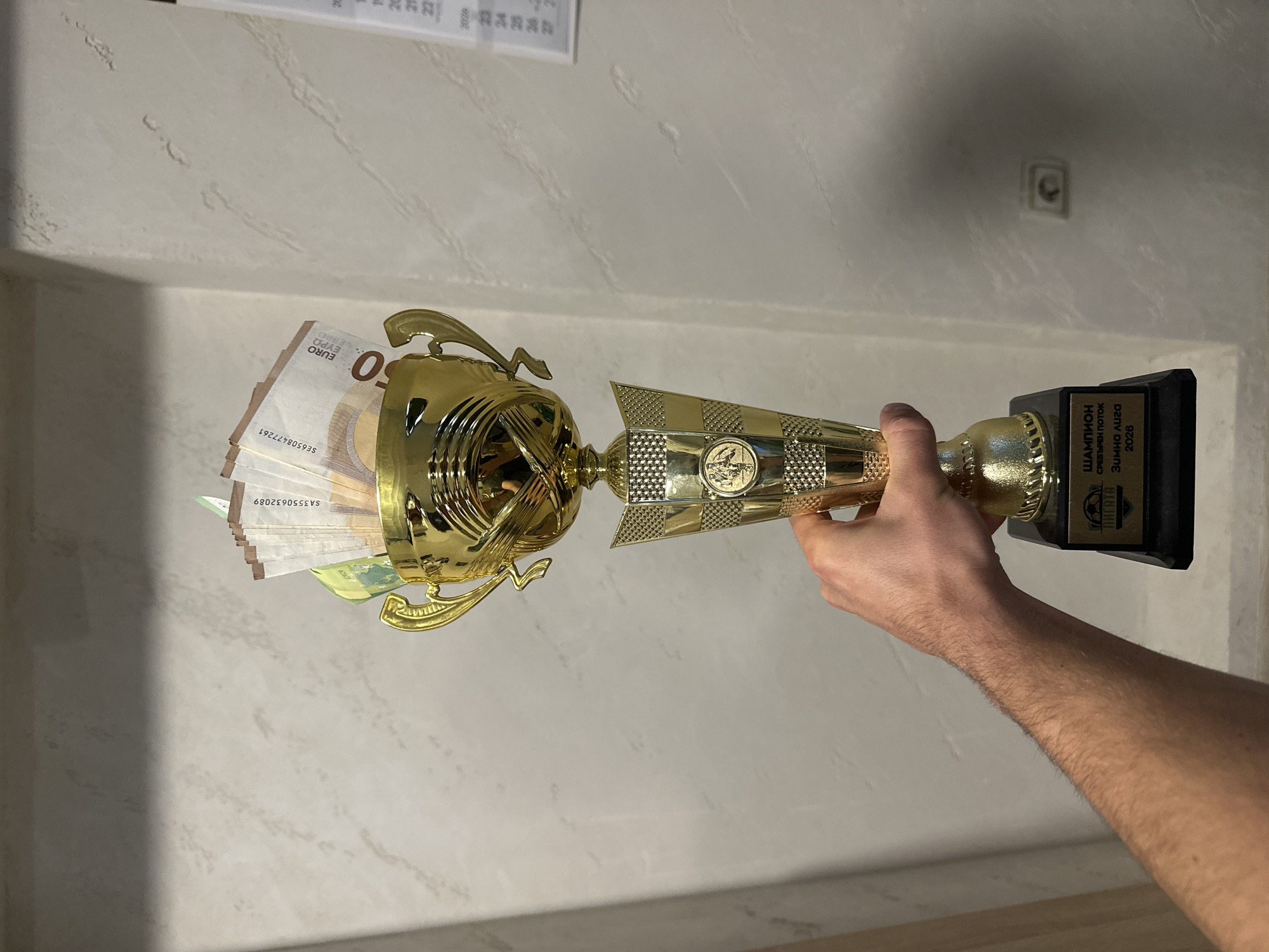 A person's hand firmly holds a golden trophy filled with Euro banknotes, set against a plain, light-colored textured wall in an indoor setting, likely in Ivailo, Bulgaria.

The central subject is a shiny, gold-colored trophy with a black rectangular base, held by a light-skinned, hairy forearm and hand. The trophy's body features intricate patterns, including a checkerboard design and a circular emblem on its mid-section. The cup-shaped top of the trophy is visibly filled with fanned-out 50 Euro banknotes. Several notes are clearly visible, displaying "50 EURO EYPQ EBPO" and serial numbers such as "SA3550632089" and "SE6508477261".

The black base of the trophy bears an inscription in Cyrillic text. The text reads: "ШАМПИОН" (CHAMPION), "СРЕБЪРЕН ПОТОК" (SILVER STREAM), "Зимна лига" (Winter League), and the year "2026". Below the text, a logo resembling a stylized football (soccer ball) is present. This inscription indicates the trophy is an award for winning a "Winter League" championship, with "Silver Stream" likely being a team or league name. The year 2026 suggests a future event or an unusual inscription.

In the background, a simple, textured white or off-white wall is visible. A light switch or electrical outlet is mounted on the wall in the upper right. A small portion of what appears to be a calendar, showing some numbers and the year "2026", is seen in the top left corner. The overall lighting suggests an indoor environment, and the trophy filled with cash signifies an achievement and a substantial prize.