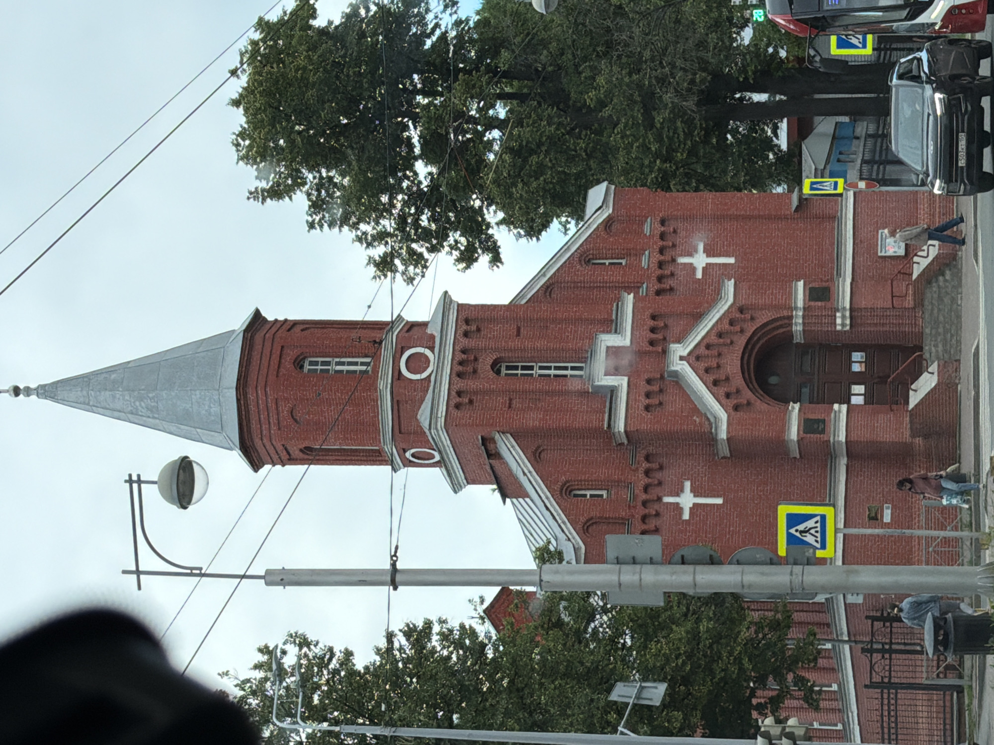 The image depicts a red brick church with a tall, grey, pointed spire. The architecture features decorative white trim around the windows and roofline, and a white cross is visible on the church's facade. A person in a pink jacket and blue jeans is walking up a set of steps leading to the church entrance. Next to the steps is a yellow diamond-shaped road sign with a blue pedestrian symbol. In the background, trees, a street light, and cars are visible. One car has a license plate with Cyrillic characters. The sky is overcast, suggesting it is daytime. The scene appears to be in a town or city. There is a sign on the building with some text that is not legible. Another yellow sign with a pedestrian symbol is visible on a pole.