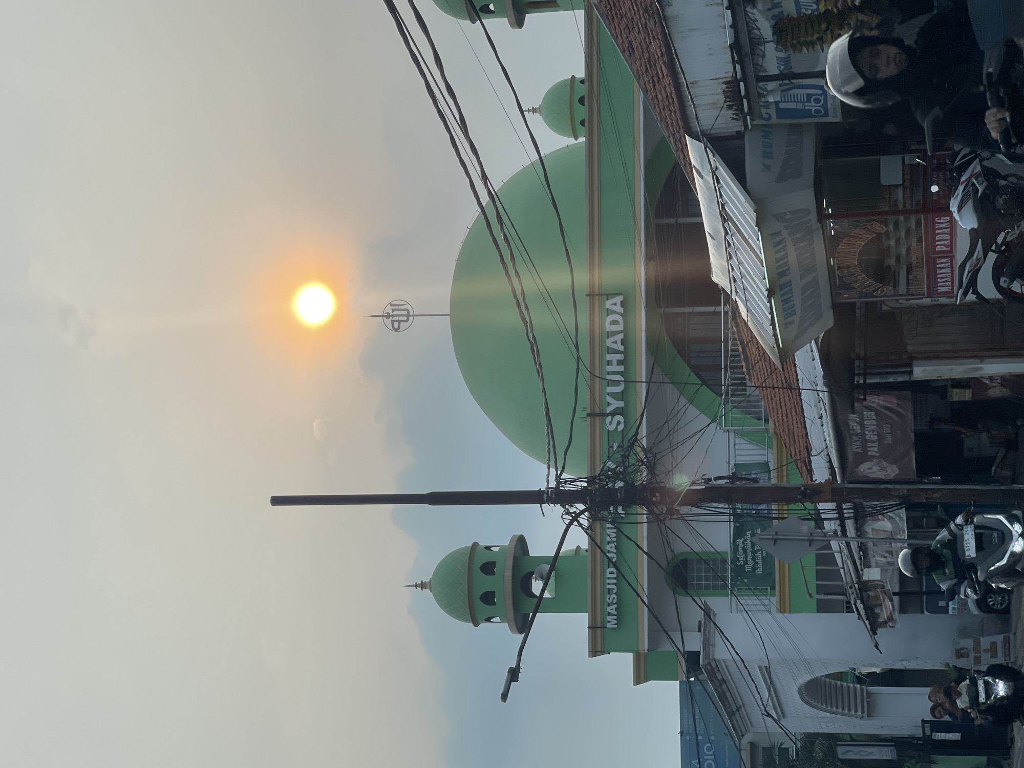 The image depicts the "Masjid Jami' Syuhada" mosque in South Tangerang, Indonesia, during what appears to be sunset. The mosque features a prominent green dome and a minaret, both characteristic of Islamic architecture. The sun, a bright orb with visible rays, is setting in the hazy sky, casting a warm glow on the scene. In the foreground, a street scene unfolds with several motorcycles, some with riders, navigating the road. Overhead, a tangle of electrical wires crisscrosses the frame, a common sight in urban environments. Buildings line the street, including what appears to be small businesses or shops. The overall atmosphere is that of a bustling urban environment with a prominent religious structure, set against the backdrop of a dramatic sunset.
