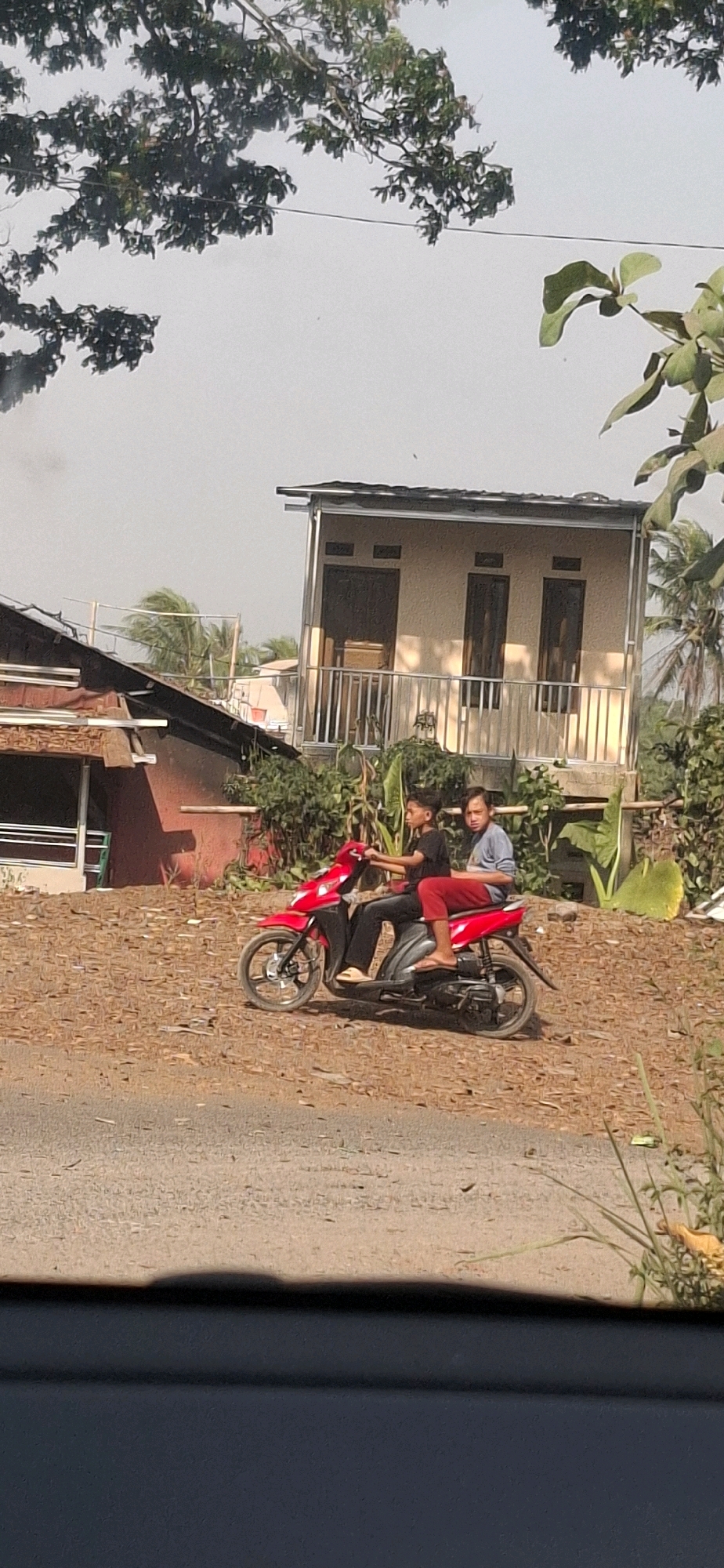 In Cibinong Hilir, Indonesia, two young males are riding a red scooter on a dirt road. The driver, wearing a black t-shirt and dark pants, operates the vehicle, while the passenger, in a grey long-sleeved shirt and red pants, sits behind him. They are positioned in the middle ground of the image. To the left, a red-colored building with a white trim is partially visible, and further back, a modern, light-colored building with three windows and a metal railing is seen. The environment is characterized by dry, brown foliage on the ground and scattered green vegetation. Trees with visible branches and leaves frame the upper portion of the scene, and a hazy sky is overhead. The foreground features a grey asphalt surface, suggesting the edge of a road.