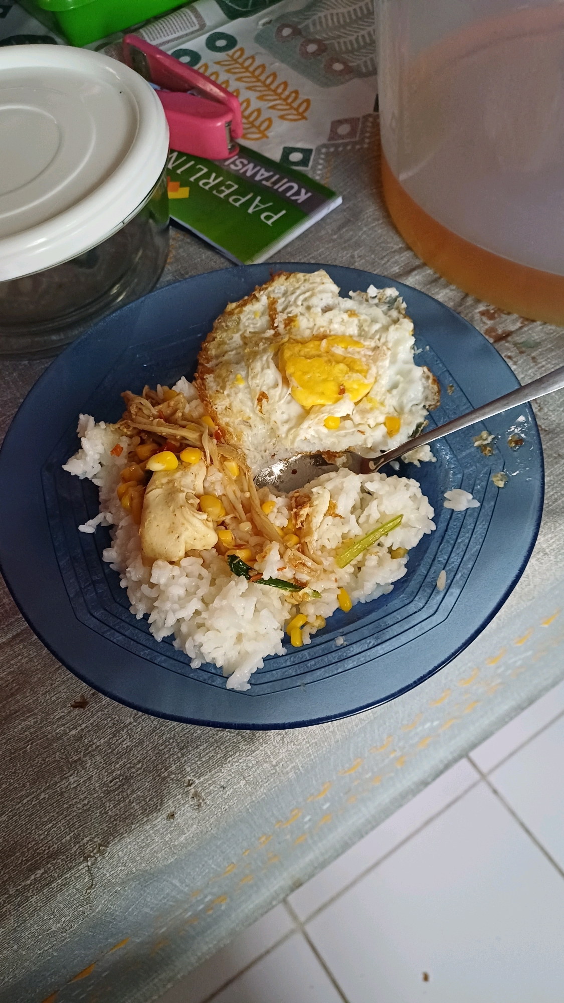A plate of food, consisting of white rice, a fried egg, and some mixed vegetables, is placed on a patterned tablecloth. The food is centered in the frame, with a fork resting on the plate. In the background, a clear plastic jug containing a light orange liquid is visible, along with a green notebook labeled "PAPERLIN KUITANSI" and a pink stapler. The scene appears to be an indoor setting, possibly a dining area in Gunung Putri, Indonesia.