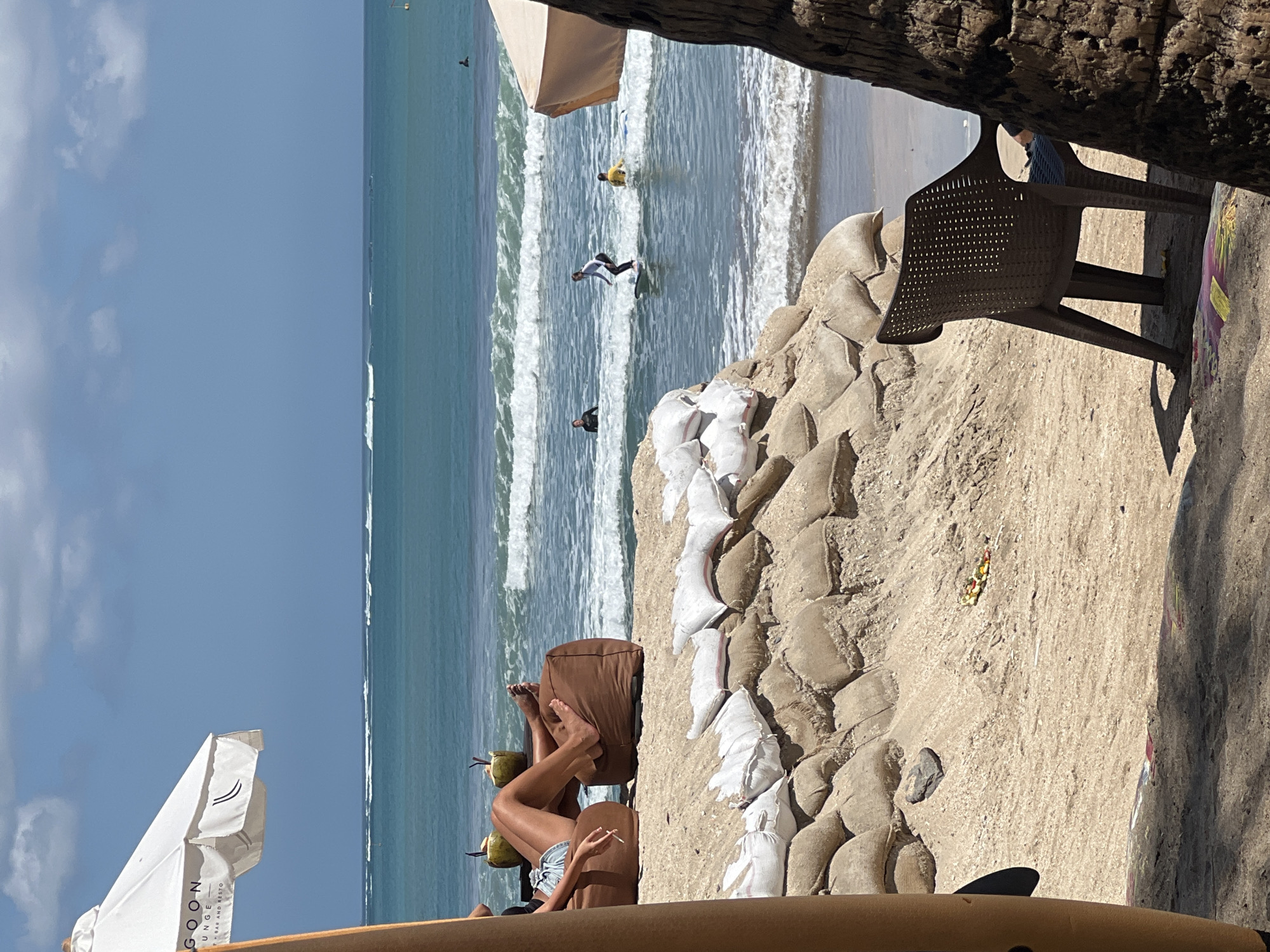 The image captures a sunny day at a beach in Kuta, Indonesia. In the foreground, a brown plastic chair sits on the sand, partially obscured by a palm tree trunk. Behind it, a sandbag barrier is piled high, with some white bags visible on top. Further back, two large brown beanbag chairs are occupied by a person lounging with their legs extended, holding a cigarette and a drink. Two coconuts with straws are placed next to them. Beyond the beach, the turquoise ocean stretches to the horizon, with several people surfing on gentle waves. A white beach umbrella with the word "GOON" and "LOUNGE" printed on it is visible on the left side of the frame. The sky is blue with scattered white clouds.