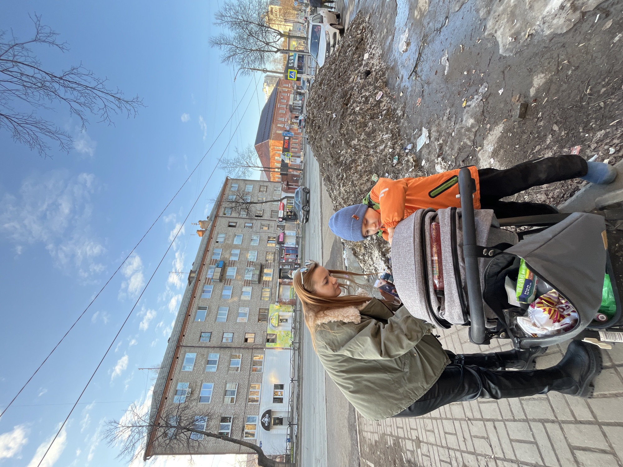 A street scene in Chaikovsky, Russia, features a woman and a child with a stroller standing on a tiled sidewalk. The woman, dressed in an olive green jacket with a fur-lined collar, black pants, and black boots, appears to be looking at something near the stroller. The child, wearing an orange jacket and blue hat, is leaning on the stroller. The stroller contains various items in its storage compartment, including packaged snacks.
Behind them, a multi-story brick building with numerous windows stands along the street. A pile of dirty snow sits on the edge of the sidewalk. Other buildings, trees, and vehicles can be seen further down the street. The sky is blue with scattered clouds.