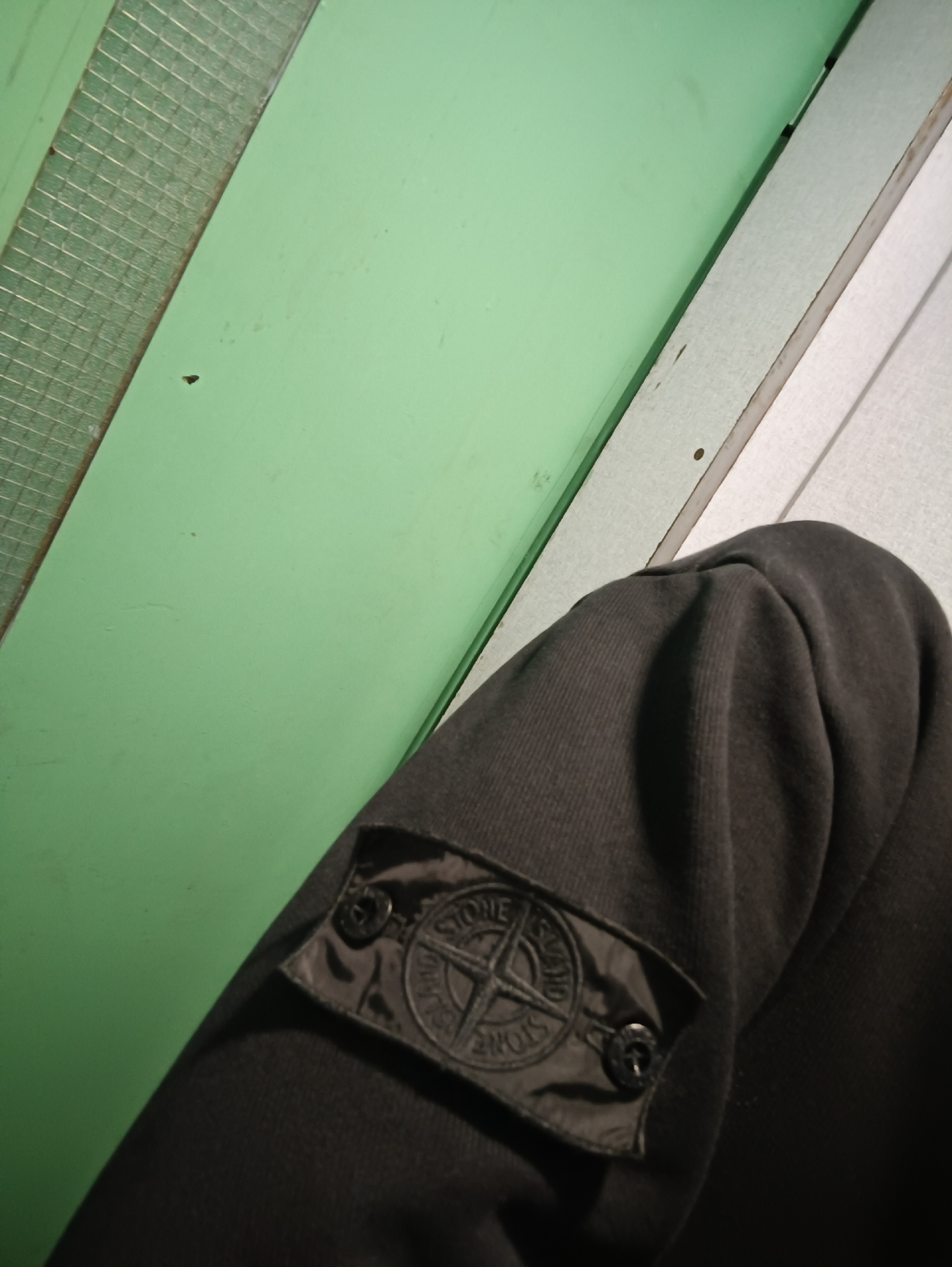 The image displays a close-up shot of a person's left arm, wearing a dark, possibly black or dark grey, long-sleeved garment. A black, rectangular patch featuring the Stone Island compass logo is prominently visible on the upper sleeve, secured by two small black buttons. The brand name "STONE ISLAND" is inscribed in a circular pattern around the central compass motif on the patch.
The background comprises distinct vertical surfaces. To the left, there's a light green wall or panel, with a portion on the far left edge exhibiting a grid-like or corrugated texture. To the right of the green surface, a lighter, possibly white or off-white, wooden or panelled surface with visible vertical lines is present. The overall setting appears to be indoors or within a semi-enclosed environment, with artificial lighting illuminating the scene. The image is contextualized as being taken in Sofia, Bulgaria. No specific activity is depicted, apart from the presentation of the garment. Emotions are not discernible from this partial view of the subject.