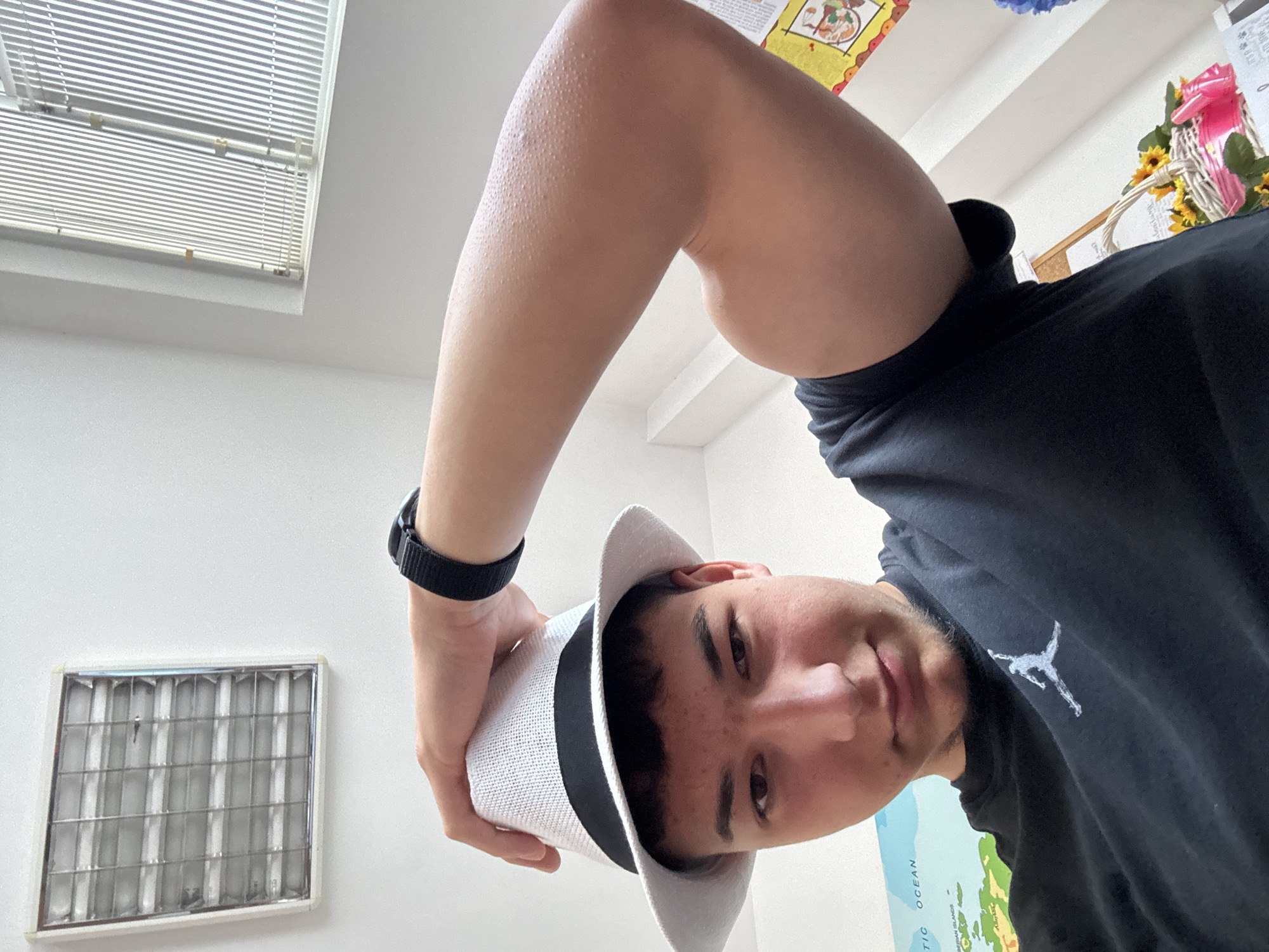 A close-up, low-angle selfie captures a young man holding a white fedora-style hat. The image is tilted, presenting the man's face in the lower center, looking directly at the camera with a neutral to slightly smiling expression. He has short, dark hair and subtle facial hair. Visible goosebumps cover his exposed arm, which is raised and bent at the elbow.
He is wearing a black t-shirt featuring a white "Jumpman" logo, identifying it as the Jordan brand. A black-strapped wristwatch is visible on his left wrist. The hat he holds is white with a distinct black band.
The background reveals an indoor setting with white walls. In the upper left corner, a window fitted with horizontal blinds allows natural light into the room. Below the window, a rectangular fluorescent light fixture with a grid cover is mounted on the wall. The upper right portion of the background includes wall decorations, notably a woven basket containing artificial sunflowers and a pink bow, and possibly other craft items. A section of a world map is visible in the lower right, with the words "ATLANTIC OCEAN" and "CARIBBEAN ISLANDS" partially legible. The lighting is bright and consistent, suggesting daytime. The location context is Zlatograd, Bulgaria.