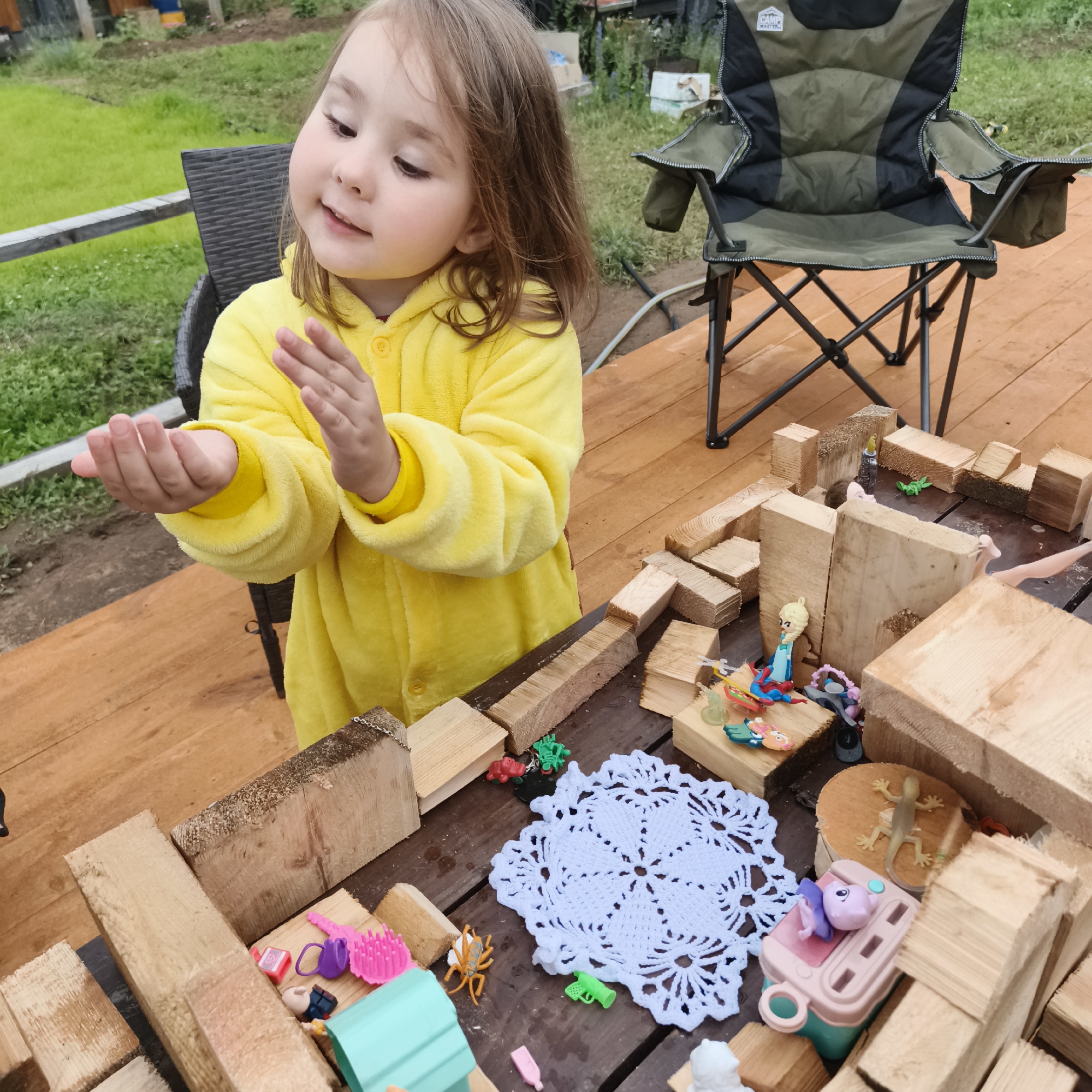 A young girl in a yellow onesie is playing with wooden blocks and small toys on a wooden deck. The girl is sitting on the deck, and her hands are outstretched. She is looking down at the blocks and toys with an expression of concentration. The toys include several small plastic animals, a toy watering can, a pink hairbrush, a small doll, and what appears to be a toy medical device. The blocks are arranged to form a structure, possibly a fort or a house. In the background, there is a green lawn, a fence, and a folding camping chair. The lighting suggests it is daytime, possibly overcast given the soft shadows. The setting appears to be outdoors, likely a backyard or patio. The context provided indicates the location is Chaikovsky, Russia.