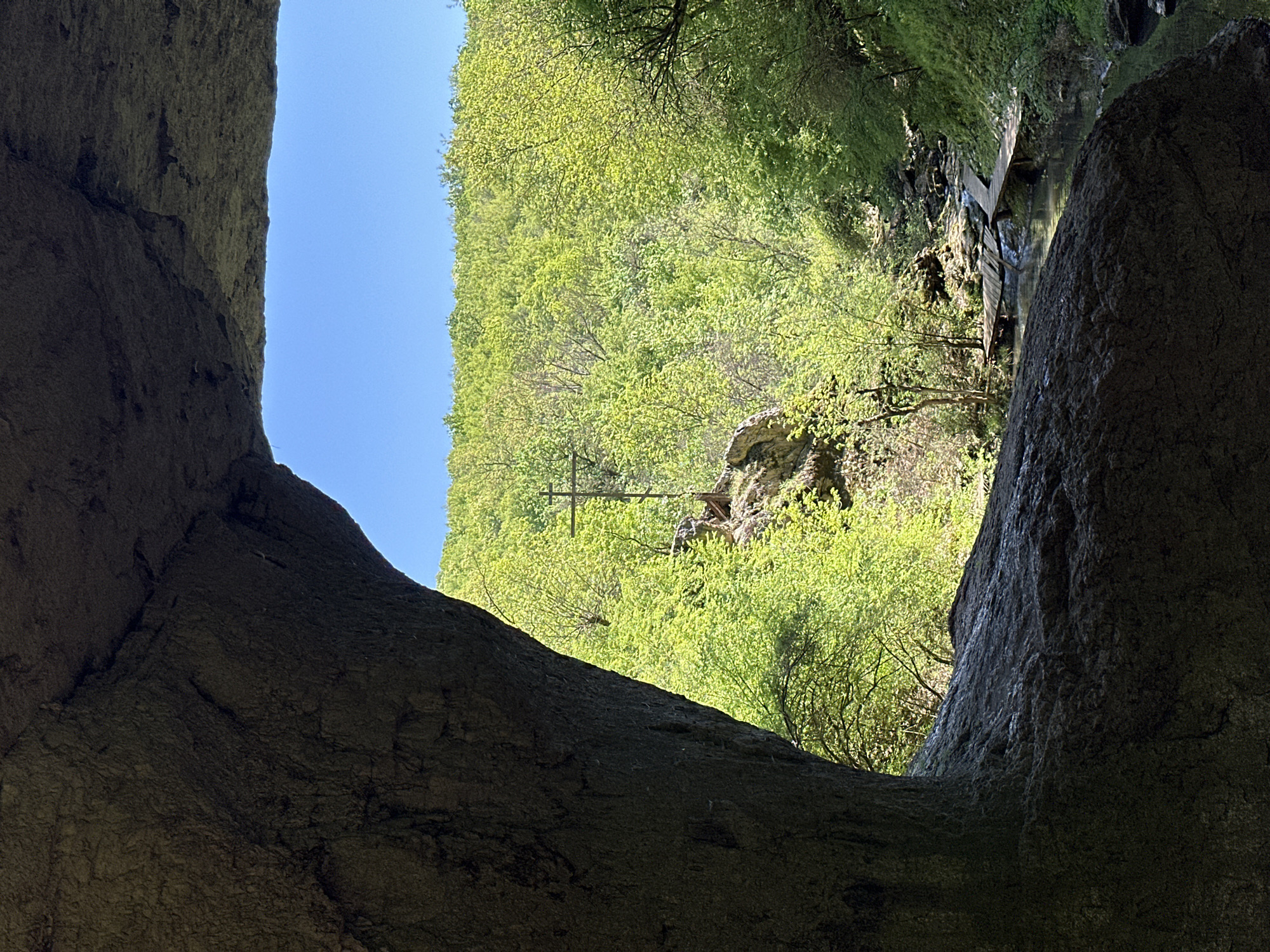 The image depicts a view of a gorge landscape, possibly in the Lilyache area of Bulgaria, seen from within a dark, enclosed space that might be a cave or tunnel. The foreground is dominated by rough, dark rock formations, creating a frame around the view. Beyond the rock, a bright, green landscape is visible, with dense foliage covering the slopes. A blue sky is visible in the top left corner. A cross-like structure is faintly seen against the green backdrop. In the middle distance, a wooden structure, potentially a bridge, spans a narrow, shaded section of the gorge.