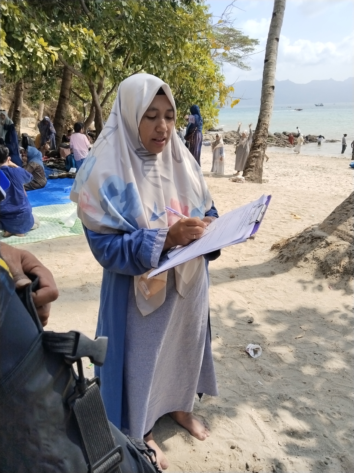 A woman wearing a light-colored hijab with floral accents and a blue dress stands on a sandy beach in Prigi, Indonesia, holding a clipboard and a pen. She is looking down at the clipboard and appears to be writing or taking notes. Behind her, other people are visible on the beach and in the water, some sitting on mats, others wading. Trees line the back of the beach, and the ocean stretches out to the horizon with distant hills. A coconut tree trunk is prominent in the foreground to the right. The scene suggests a casual gathering or event by the sea.