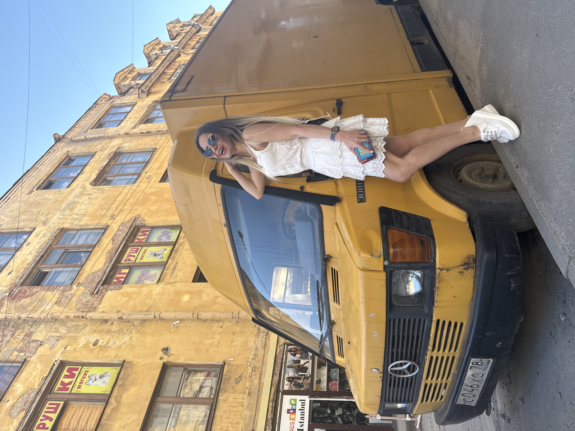 A woman in a white lace dress and white platform sneakers poses next to a yellow Mercedes-Benz van on a city street in Saint Petersburg, Russia. The van's license plate reads "C046XC 78 RUS". Behind the van, a weathered yellow building with multiple windows and peeling paint is visible. Signs with Cyrillic text and images of animals are displayed in some of the windows. To the left of the van, a storefront with the words "Istanbul Bags Shop" is partially visible. The woman is wearing sunglasses and holding a smartphone. The sky is clear and blue, and power lines are strung across the top of the frame. The street is paved with asphalt and bordered by a concrete curb.