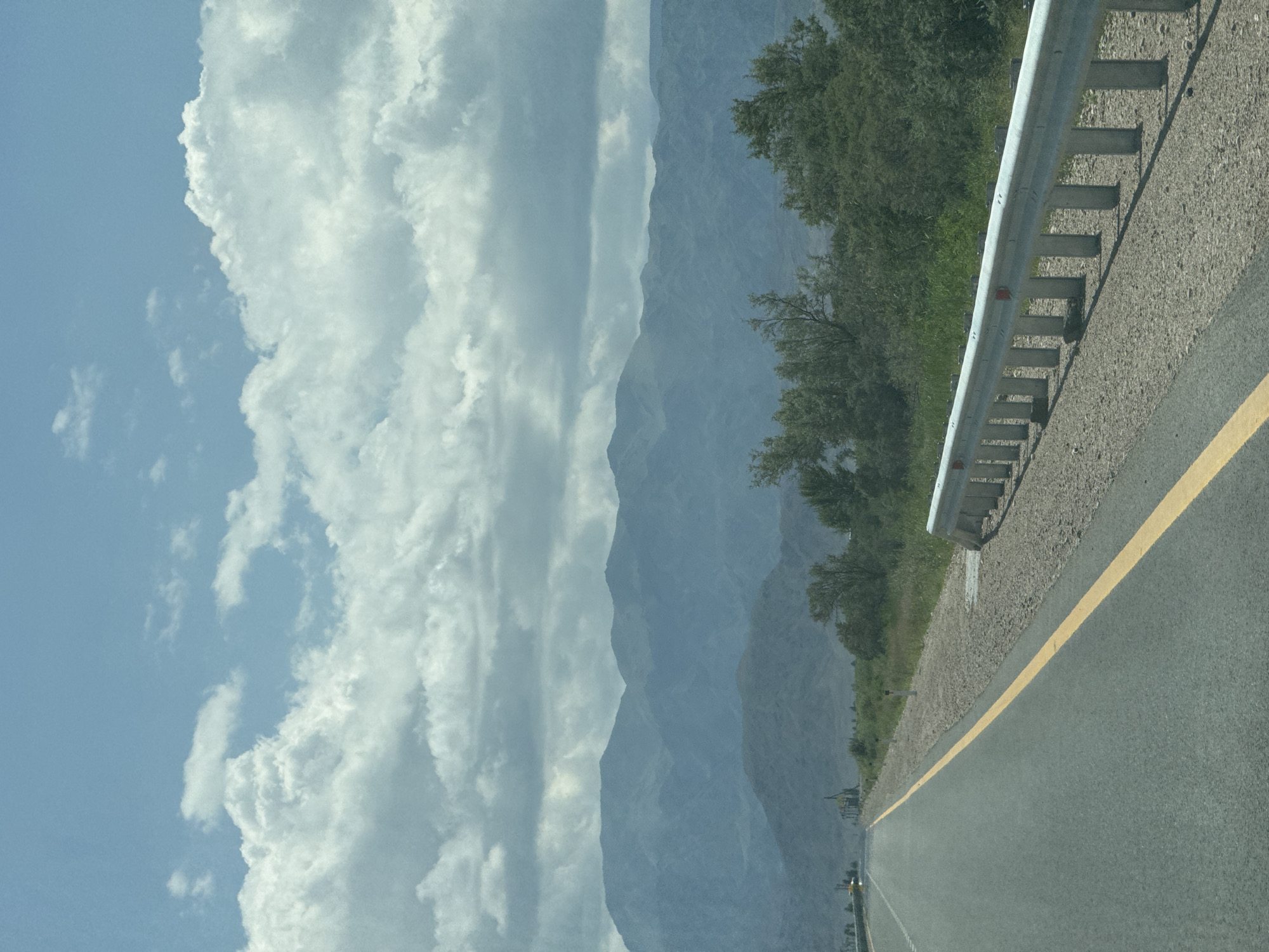The image shows a roadside view with a highway stretching into the distance. The highway has a yellow line marking the lane. On the right side of the road, there is a guardrail made of metal. Beyond the guardrail, there is a strip of gravel and then vegetation consisting of bushes and trees. In the background, there are mountains covered in a hazy blue. The sky above is blue with scattered white clouds. There are no people or animals visible in the image. The scene appears to be a rural or semi-rural area. The location is described as being in the city of Balykchy, Kyrgyzstan.