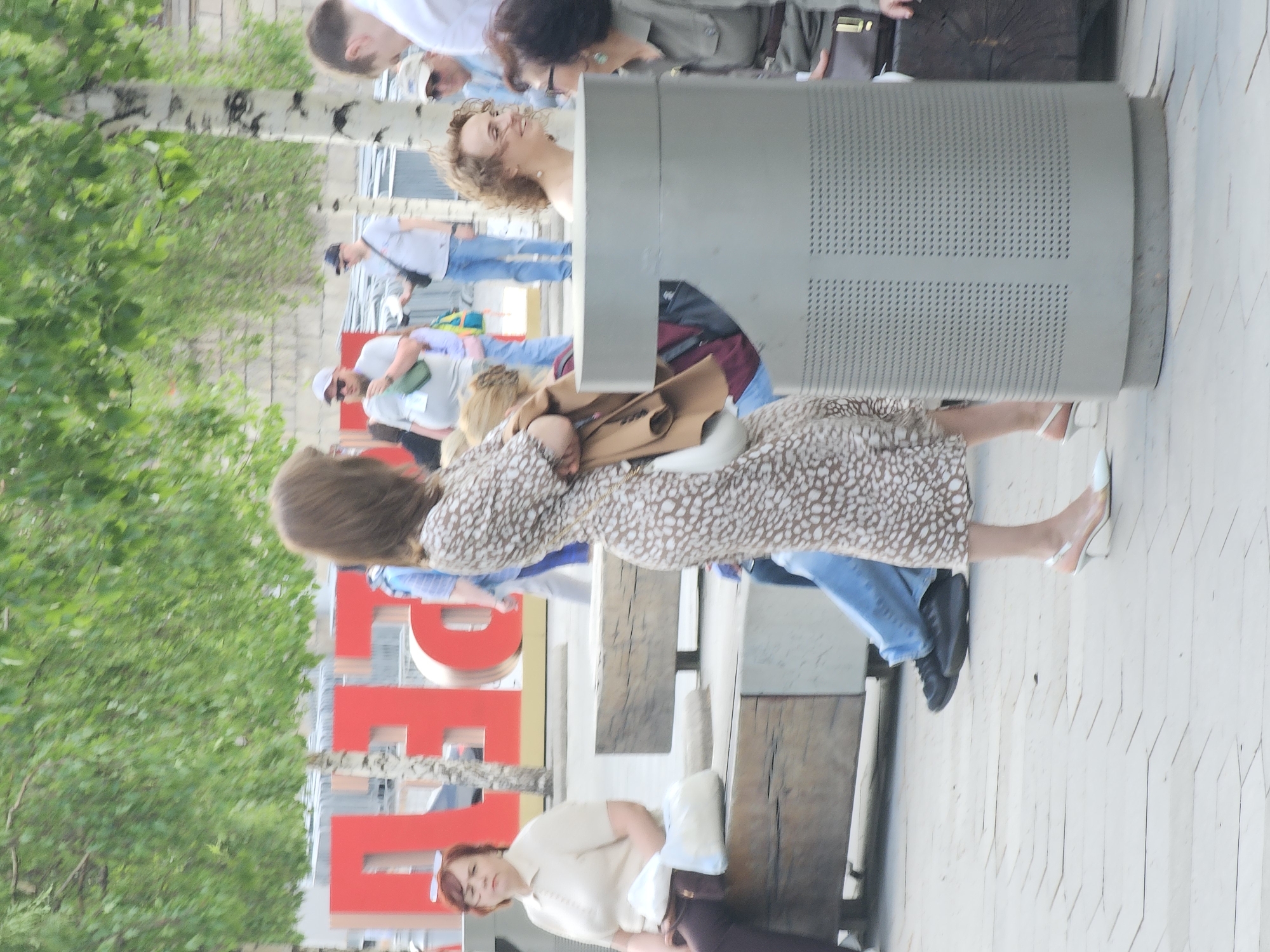 The image depicts an outdoor scene with several people and objects. A woman in a brown patterned dress and white heels stands near a large gray metal structure. In the background, there are several other individuals, including a woman sitting on a bench with a white pillow. The word "RED" in large red letters is visible near a tree. The scene appears to be in Moscow, Russia, as the objects and environment point towards this location.