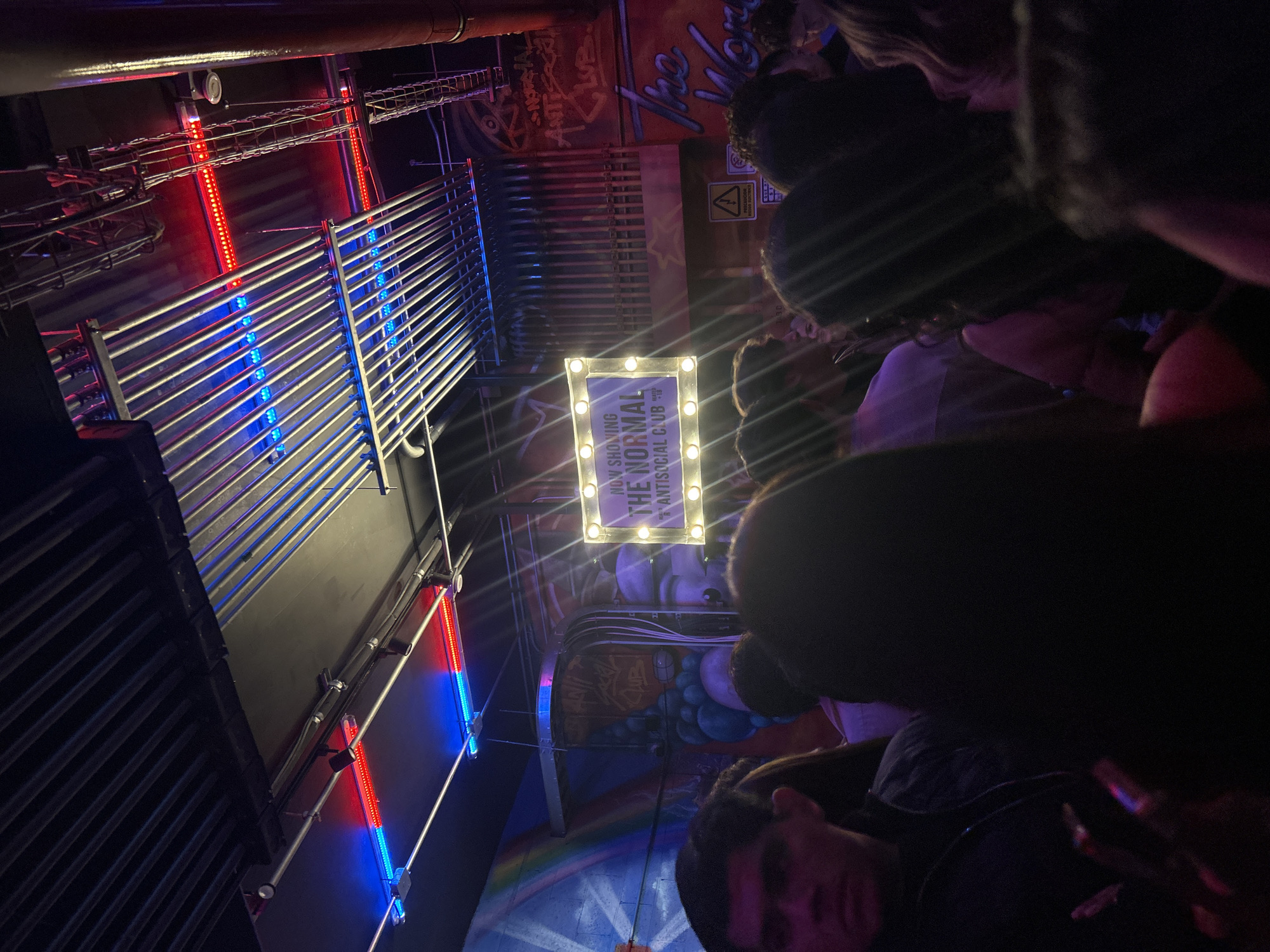 Captured at night, this image shows an indoor scene, possibly a club or bar in San Bernardino Tlaxcalancingo, Mexico. The focus is on a sign illuminated by lights that reads "NOW SHOWING THE NORMAL ANTISOCIAL CLUB." Around the sign and in the foreground, there are numerous indistinct figures of people, seemingly an audience or crowd. The environment includes exposed ductwork on the ceiling, accented with blue and red lighting. A mural adorns the wall behind the sign.