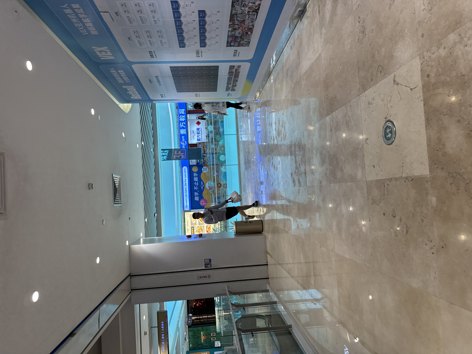 The image shows an interior shot of a brightly lit shopping mall, likely in Haizhu District, China, given the presence of Chinese signage. The floor is made of polished beige marble tiles, reflecting the overhead lights and ceiling. A large blue and white promotional banner, featuring Chinese text and images of robots, dominates the right side of the frame. The banner appears to advertise a robotics competition or educational program, with text such as "VEX 亚洲机器人锦标赛教学成果" visible.
In the mid-ground, two individuals are visible. On the left, a man wearing a grey hooded sweatshirt and black shorts walks away from the camera, holding a white plastic bag in his right hand and looking at his phone. He is near a metallic trash can. On the right, a woman in a white patterned shirt and black pants walks towards the left of the frame, also holding a white bag.
The background features more mall architecture, including glass railings, doorways, and what appear to be shop entrances with illuminated displays and signs, some in Chinese characters. A bright blue lighting scheme is evident in the background, suggesting a modern retail environment. A circular floor fixture with green arrows pointing up and down is visible in the foreground, indicating a potential directional marker or emergency exit indicator. The overall atmosphere is clean, modern, and commercial.