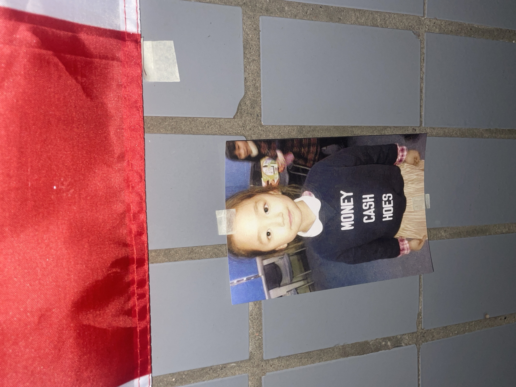 A printed photograph of a young girl is prominently displayed, taped onto a grey tiled surface, possibly a sidewalk or wall. To the left, a portion of a red and white fabric flag is visible, partially obscured by the photograph and also secured to the tiled surface.
The photograph, oriented vertically, captures a young girl with light skin and dark hair, looking directly at the viewer with a neutral to slightly pleasant expression. She is wearing a dark, long-sleeved shirt with white text stacked vertically, clearly reading "MONEY," "CASH," and "HOES." Below her shirt, a light-colored, possibly beige or light brown, skirt is visible. The background within the photograph is blurred, suggesting an indoor setting with subtle hints of other people and objects.
The photograph is held in place by two pieces of clear or frosted adhesive tape, one at the top and another near the bottom right corner. The grey tiled surface consists of rectangular tiles with darker grout lines, creating a grid pattern. The flag on the left, primarily red with a white strip along its top edge, appears to be made of textile material. The lighting in the scene suggests it is daytime. The activity depicted is the display of this photograph in an unknown, possibly public, setting within Russia.