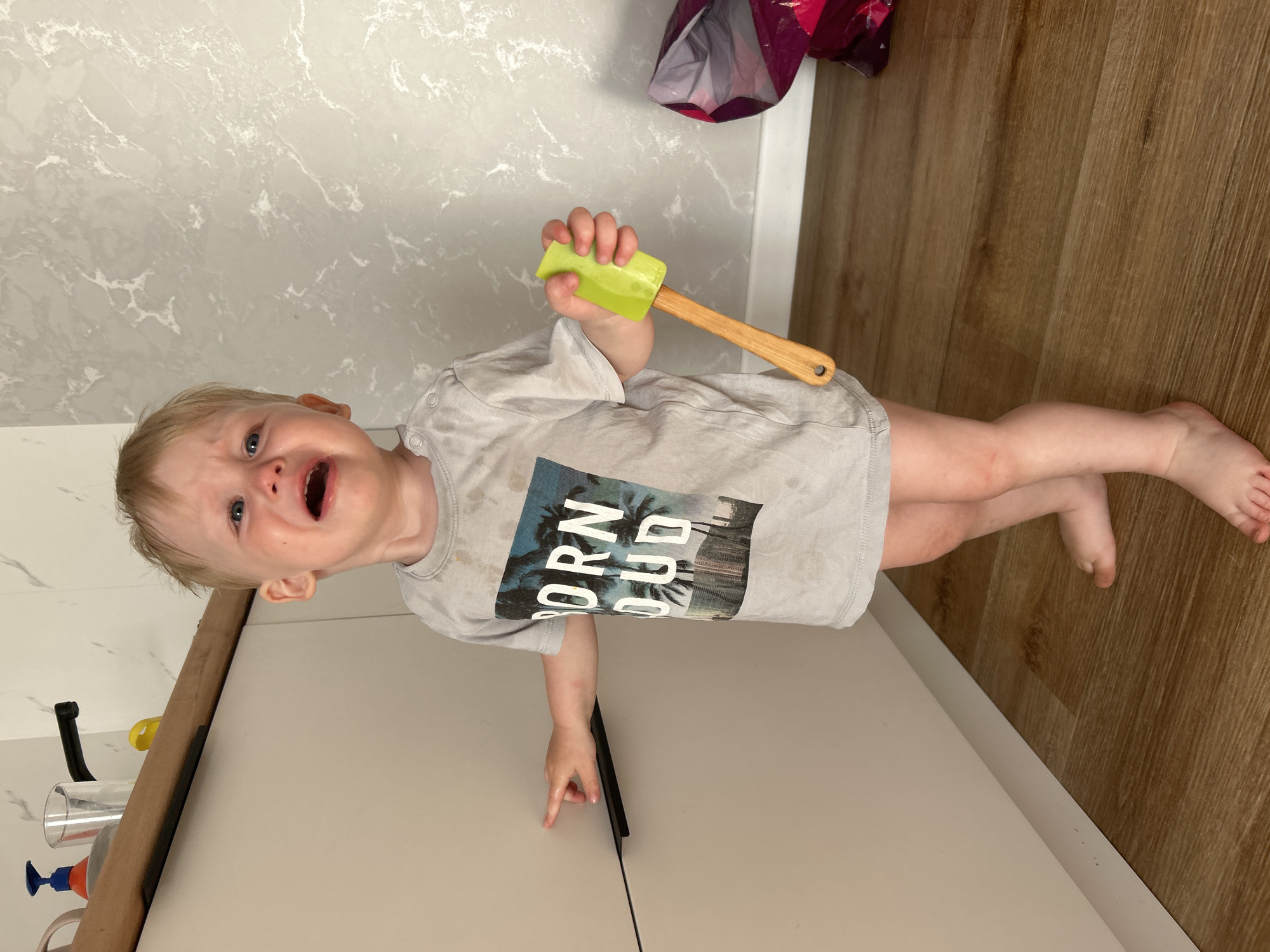 A blonde-haired toddler with blue eyes is standing in a room, crying loudly. The toddler is wearing a light gray t-shirt with a graphic of palm trees and the words "BORN LOUD". The t-shirt appears to be stained. The toddler is holding a green silicone spatula with a wooden handle in their right hand and has their left hand extended towards a light-colored cabinet. The background consists of a light-colored wall with a marble-like pattern and a wooden floor. To the right, a portion of a pink and purple patterned bag is visible. The scene appears to be indoors, possibly in a kitchen or living area, within a residential setting in Chaikovsky, Russia.