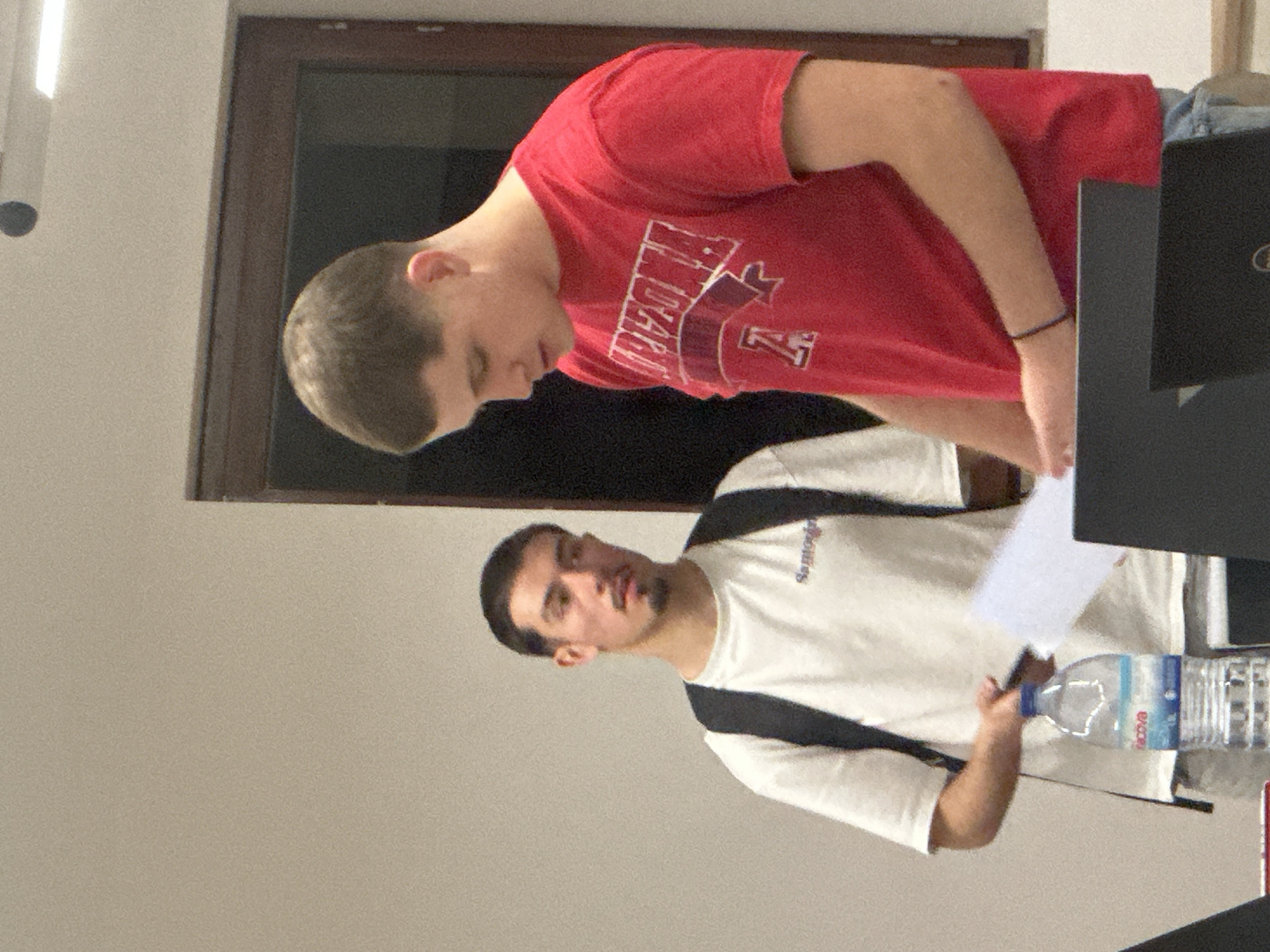The image depicts an indoor scene with two young adult males in what appears to be an educational or study environment.
In the foreground, on the right side, a male subject with short, dark hair is positioned, looking downwards. He wears a red short-sleeved t-shirt with "ARIZONA" and a letter "A" logo printed in white. A black wristband is visible on his left wrist. His lower body is partially obscured by two dark-colored laptops on a surface; one laptop features a "Dell" logo. He holds several sheets of white paper in his hands.
In the background, on the left side, a second male subject stands. He has dark hair, a mustache, and a goatee. He wears a white short-sleeved t-shirt with faint dark text across the chest (possibly "Samoglou") and a black backpack over both shoulders. He holds a dark-colored smartphone in his right hand and appears to be looking forward.
Between the two individuals, on a surface, a clear plastic water bottle with a blue label (brand "Acova") is visible. The immediate background consists of plain light-colored walls and a dark-framed window or door. Overhead, a horizontal strip light fixture and a small dark circular object are visible on the ceiling. The setting is consistent with a classroom, lecture hall, or study area within a modern educational institution, such as a university campus, located in Guimarães, Portugal. No specific landmarks are visible to identify an exact address or building within the city.