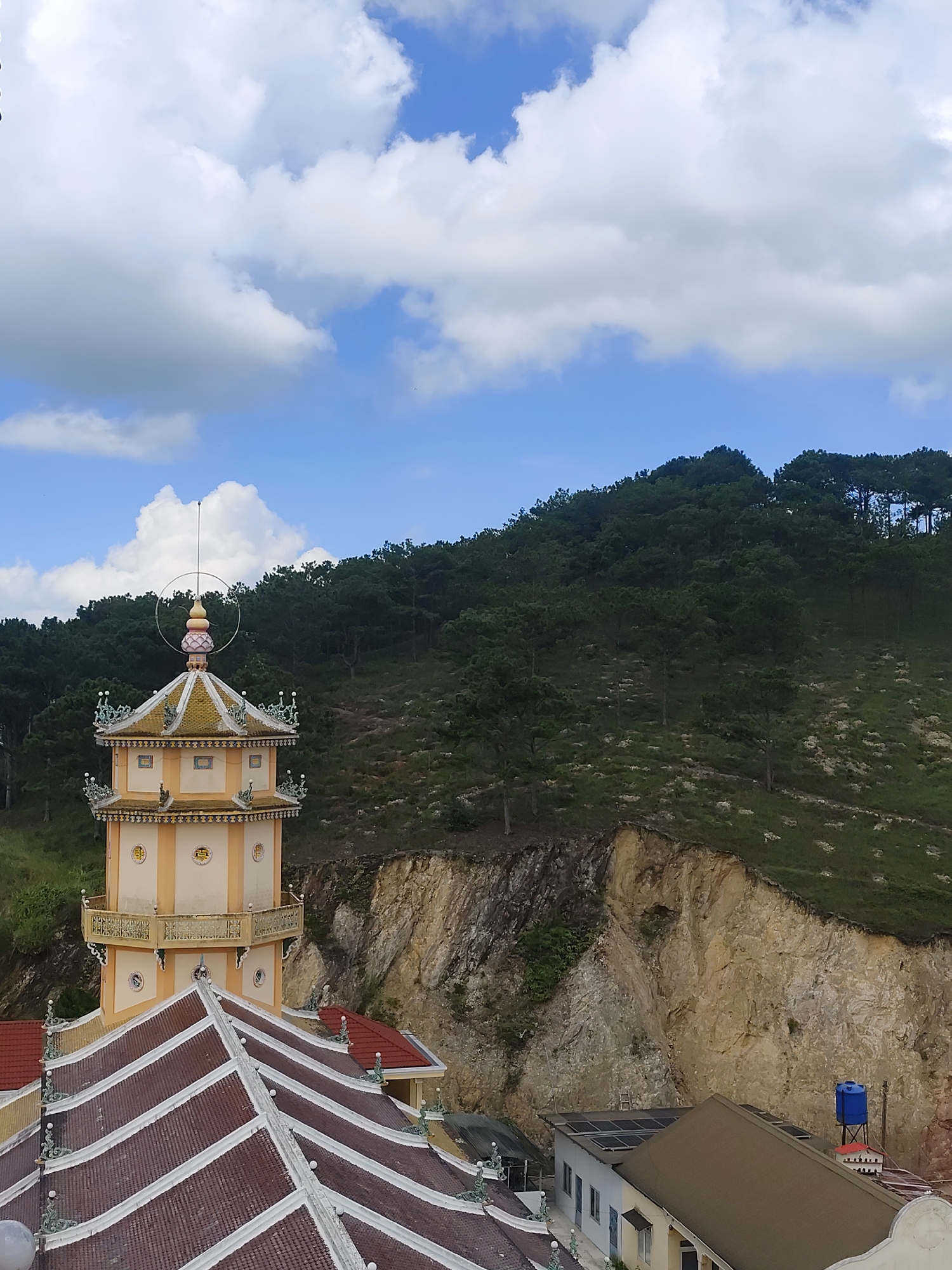The image shows a view from an elevated position, looking towards a hillside covered in dense green trees under a bright blue sky with scattered white clouds. In the foreground, a multi-tiered, ornate tower, characteristic of Buddhist temple architecture, is visible. The tower is predominantly cream and yellow, with decorative elements and a pointed roof. Adjacent to the tower, a series of reddish-brown tiled roofs with white trim and decorative finials can be seen. Further in the background, a steep, rocky cliff face is partially visible, with some greenery growing on its slopes. At the base of the cliff, a modern building with solar panels on its roof and a blue water tank are present. The overall scene suggests a temple complex situated in a natural, hilly environment, possibly in or near Lộc Quý, Vietnam, given the architectural style and vegetation. There are no people or active interactions depicted in the image.
