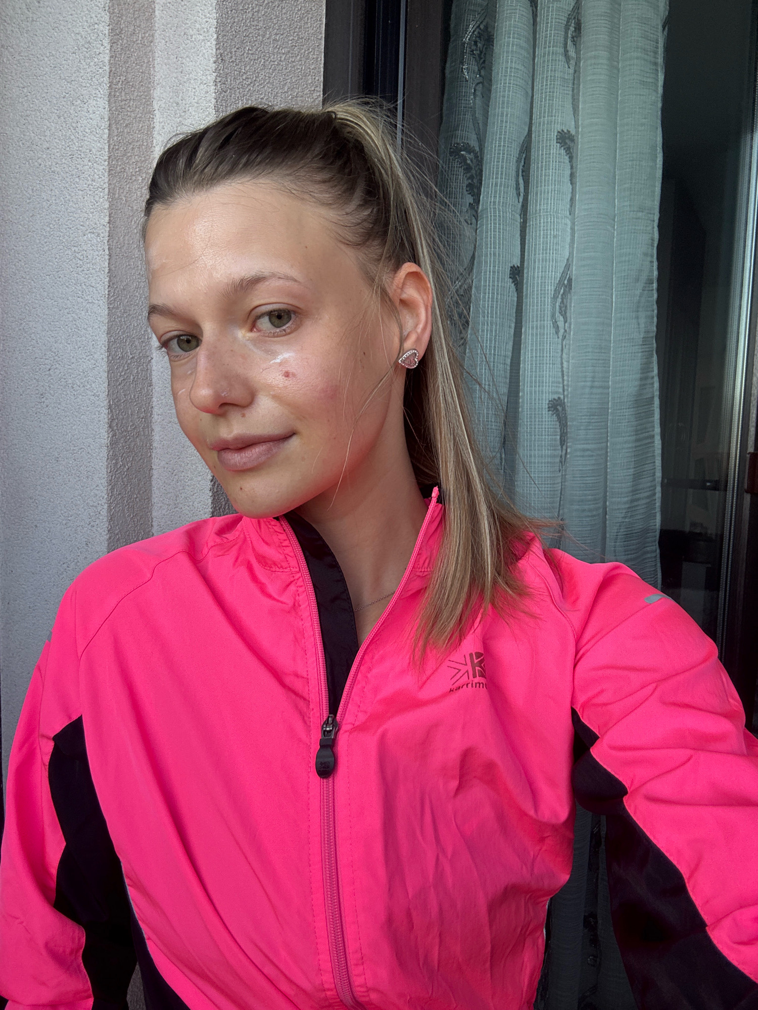 In this medium shot, a young woman with blonde hair tied in a high ponytail poses outdoors. She is wearing a bright pink and black athletic jacket. Her face is lightly made up, with a visible application of cream or concealer on her cheeks and forehead, and a small red blemish below her left eye. She is also wearing a heart-shaped earring in her left ear. The background features a textured, grey column on the left and a sheer, patterned curtain on the right, suggesting an outdoor or semi-outdoor setting, possibly a balcony or patio area. The lighting indicates it is daytime. No specific city landmarks or identifiable locations within Sofia, Bulgaria, are visible.