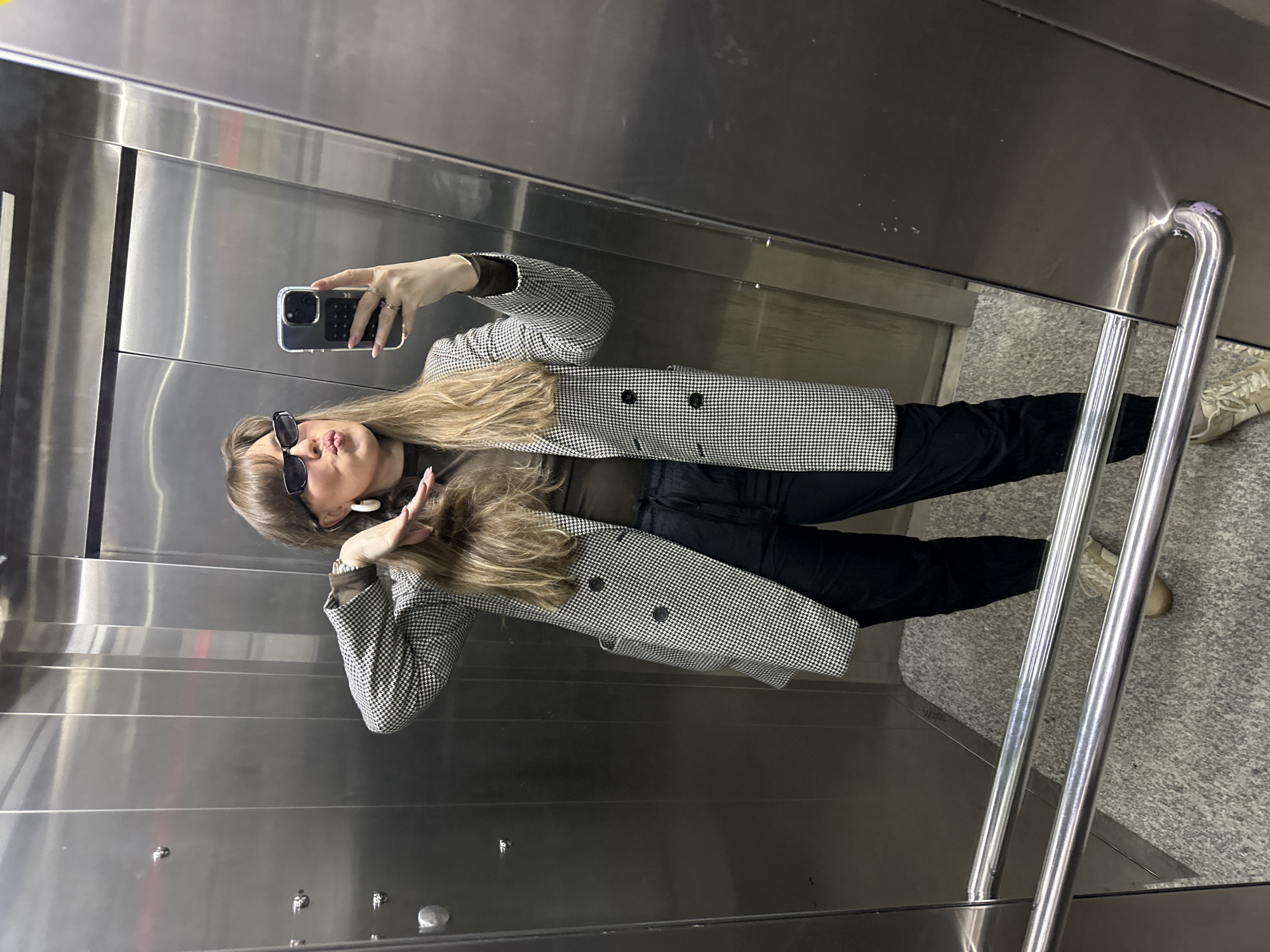A woman is taking a selfie in an elevator. She is wearing a houndstooth blazer, black pants, and sunglasses. She has long blonde hair and is holding her phone in her right hand. She is posing, making a peace sign with her left hand. The elevator has metal walls and a metal handrail. The floor is gray and speckled. The location appears to be in Tbilisi, Georgia, though this cannot be definitively confirmed from the image alone.