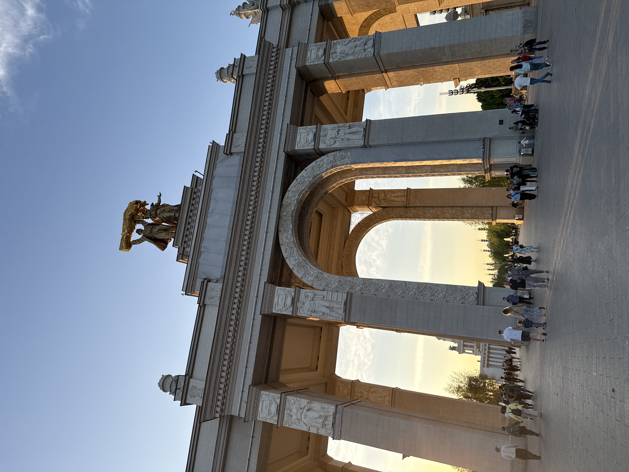 The image captures a perspective view of the Main Entrance Arch to the All-Russia Exhibition Centre in Moscow, Russia. The arch is constructed of light-colored stone with detailed ornamentation and arches. Atop the structure is a golden sculpture. Several people are seen walking through the archway, with some individuals clearly visible. The sky above is blue.