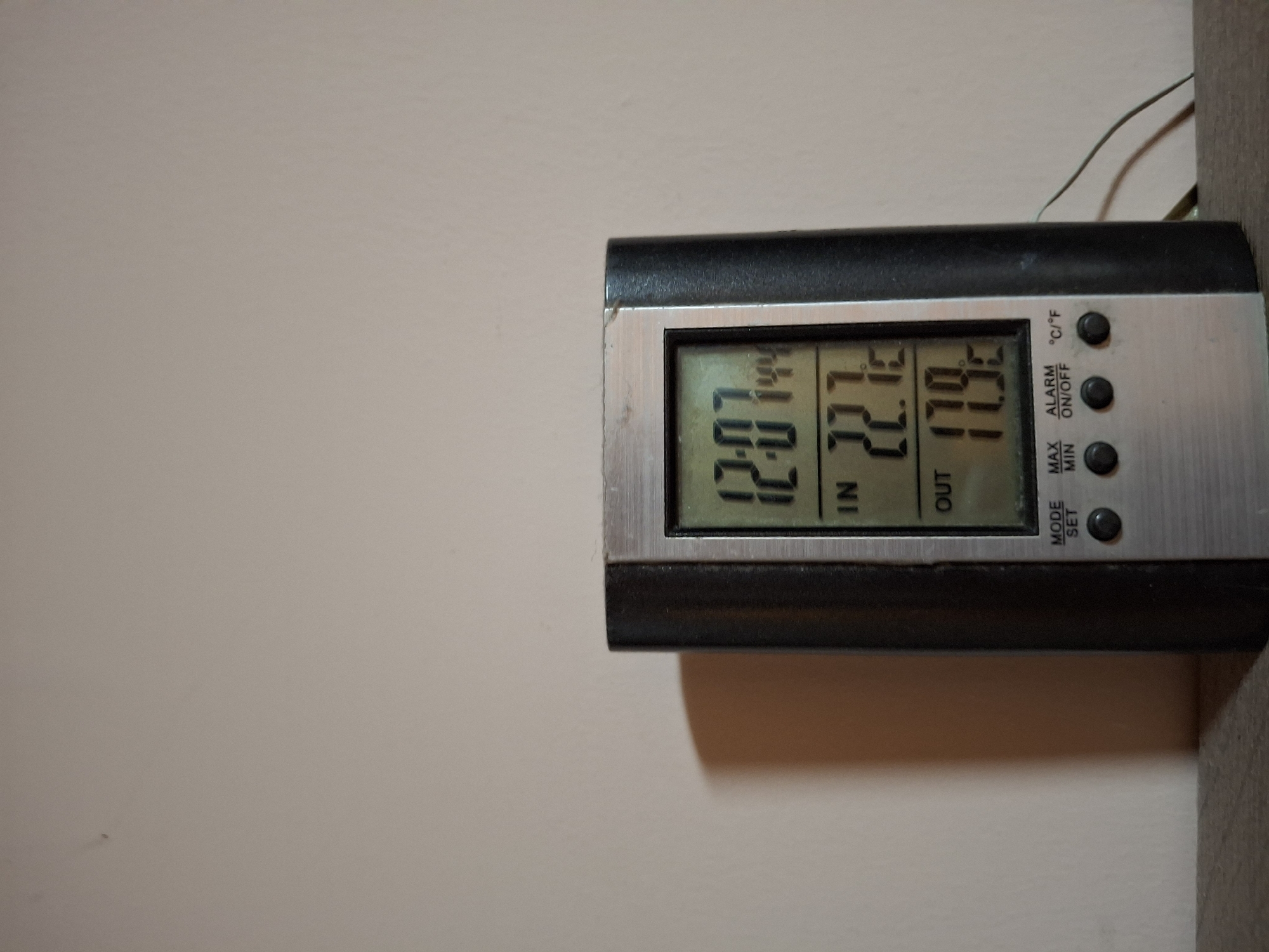 A digital thermometer is placed on a wooden surface against a plain, light-colored wall. The thermometer has a black casing with a silver front panel. The digital display shows the time as "12:07", an indoor temperature of "22.7°C", and an outdoor temperature of "17.9°C". Below the temperature readings are buttons labeled "MODE SET", "MAX MIN", and "ALARM ON/OFF", along with a "°C/°F" indicator. A thin wire extends from the right side of the thermometer, suggesting it has an outdoor probe. The lighting in the scene casts a shadow of the thermometer on the wall. There are no people or animals present. The image is taken from a medium distance, focusing on the thermometer. The background is out of focus. This scene is not specific to Ogre, Latvia; it could be any indoor location with a digital thermometer.
