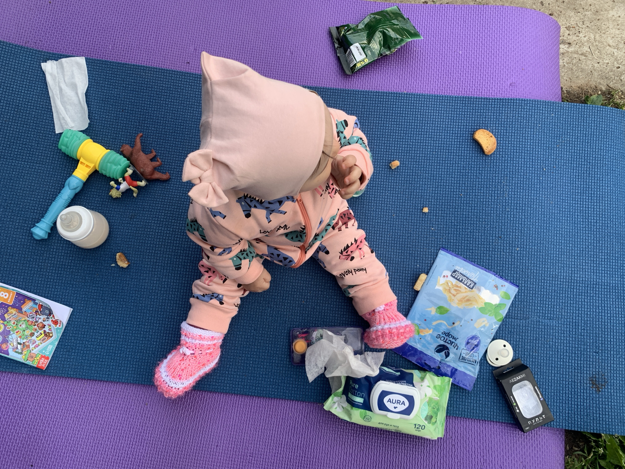 A baby wearing a pink hooded outfit and pink knitted socks is lying on a purple and blue yoga mat. Around the baby are various objects: a white tissue, a toy hammer, two animal figurines, a baby bottle, a children's book, a partially eaten snack, a small container with compartments, a packet of baby wipes, a package of fish chips, a pacifier, and a small black box labeled "AEZAKMI". The text "AURA" and "Pure Cotton" are visible on the baby wipes package. The date "18.05.2026" is printed on a green package. The text on the book reads "БУКВА" and "ЛЕНД". The words "lovely pony" are also visible on the baby's outfit. The scene appears to be set outdoors, with a concrete surface visible in the background. Since it is labeled "AEZAKMI" and has Russian on it, the location is likely to be in Chaikovsky, Russia.
