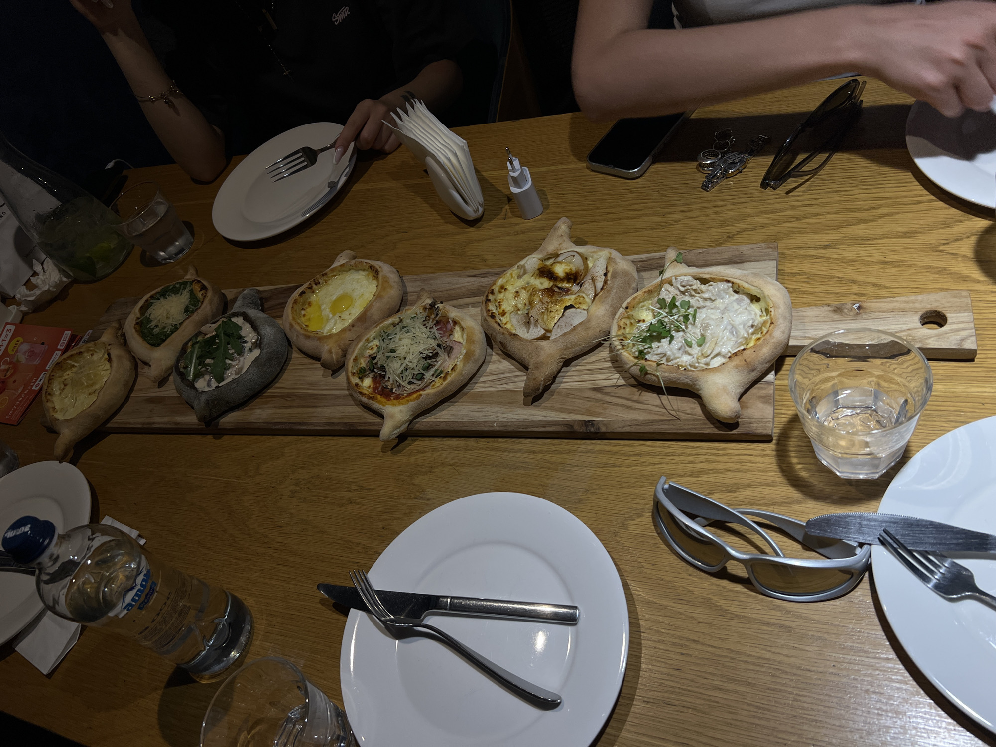 In Tbilisi, Georgia, a wooden table is laden with several delicious Khachapuri, a traditional Georgian cheese-filled bread. The Khachapuri are arranged on a wooden board, each with a unique topping, including cheese, egg, and greens. Several plates, cutlery, glasses, and a bottle of water are also present on the table, suggesting a meal in progress. A pair of sunglasses rests on the table, and a mobile phone is also visible. The scene appears to be a casual dining experience in a restaurant.