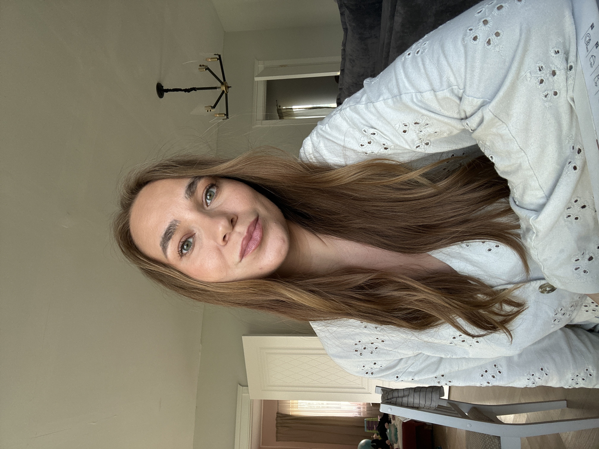 A woman with long, light brown hair and green eyes is looking at the camera, taking a selfie. She is wearing a light blue, eyelet-detailed blouse. Her face is in the foreground, and her body is angled away from the camera. The background suggests an indoor setting, possibly a home, with a chandelier visible on the ceiling and what appears to be a closet or cabinet with decorative paneling. Sunlight is coming from the left, casting soft light on her face and illuminating the room. The overall mood appears relaxed and casual. The image is rotated 90 degrees counter-clockwise.