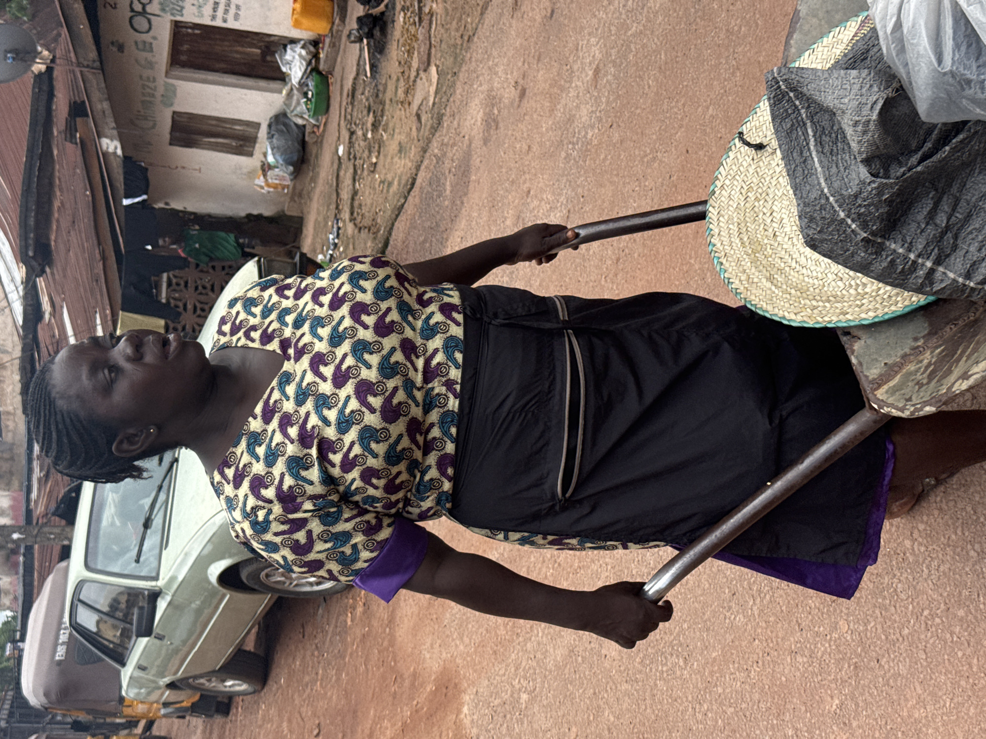 A dark-skinned woman with braided hair walks from right to left, pushing a wheelbarrow. She is wearing a yellow and purple patterned blouse and a black skirt. The wheelbarrow contains a woven fan and a black plastic bag. In the background, a light-colored sedan is parked on a dirt road. A building with a corrugated metal roof is visible on the right side of the image, with clothes hanging on a line outside. The scene appears to be a street in Enugu, Nigeria.