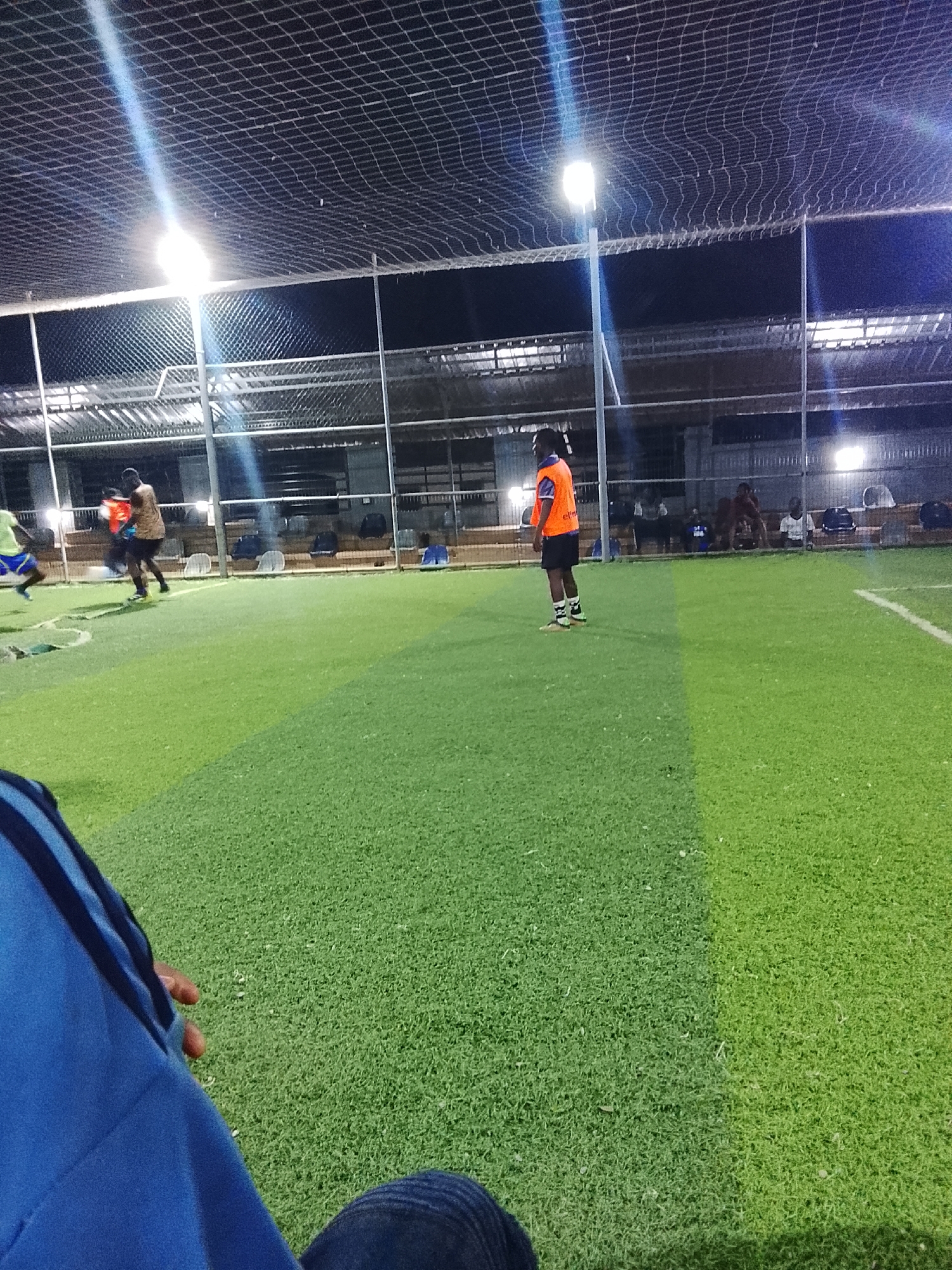 The image depicts a soccer field in Akwanga, Nigeria at night. The field has artificial turf and is surrounded by a netting enclosure under artificial lighting. Players are on the field, with some actively engaged in a game. Spectators are seated along the side. A person, partially visible, is seated in the foreground, observing.