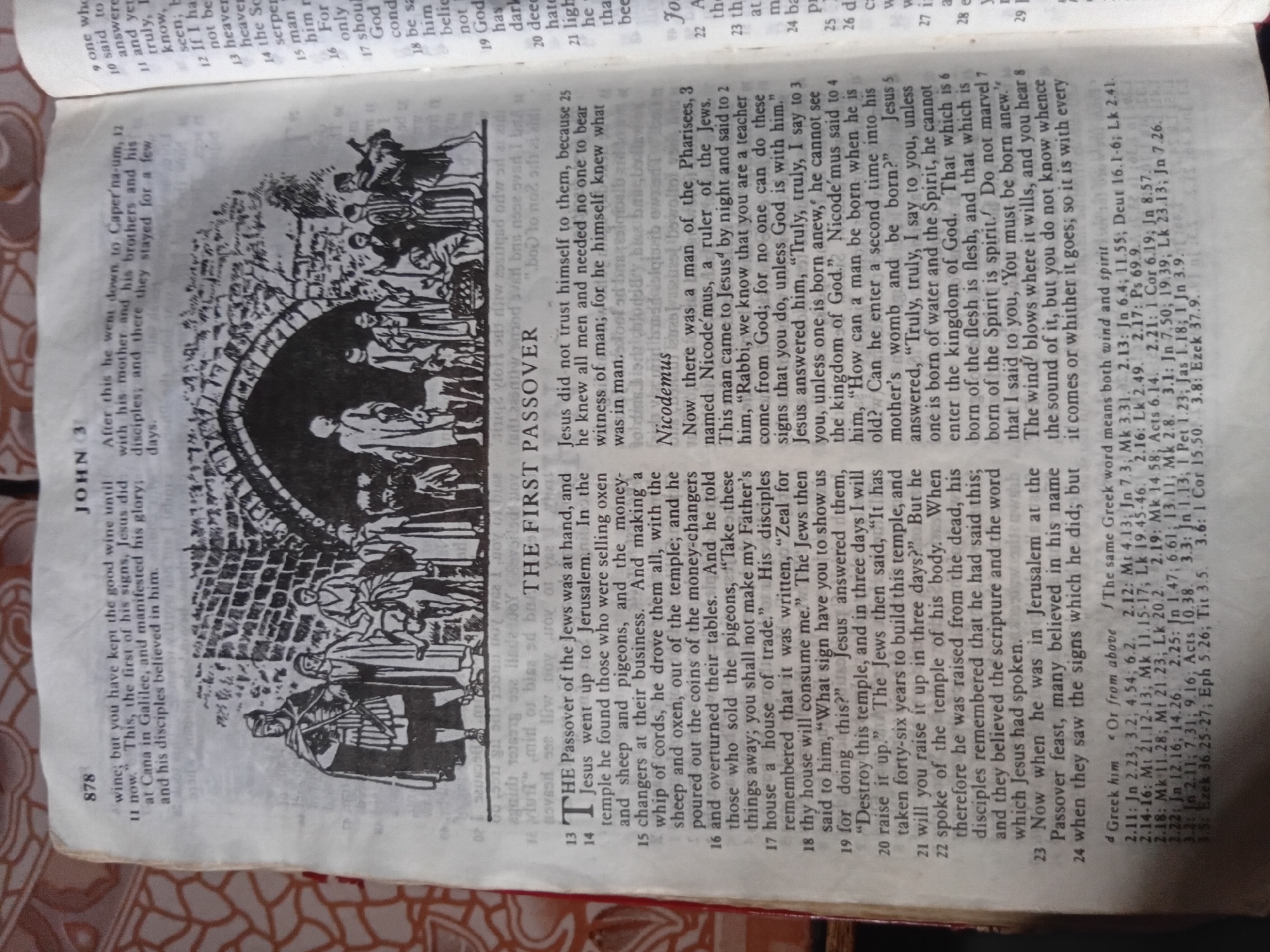 The image displays an open Bible, specifically the Book of John, Chapter 3. The page features text printed in English, accompanied by a black and white illustration. The illustration depicts a group of people in what appears to be an ancient setting, with architectural elements like arches and stone walls. Some individuals are dressed in robes, while others wear more rudimentary clothing. Animals, including sheep and a donkey, are also present in the illustration. The text describes events related to Jesus, the Passover, and a conversation with Nicodemus. The image is a close-up view of the book, with a textured, patterned surface visible in the background, suggesting the book is resting on a fabric or similar material. The lighting is consistent with indoor photography. There is no information within the image that places it in Nkpor, Nigeria.