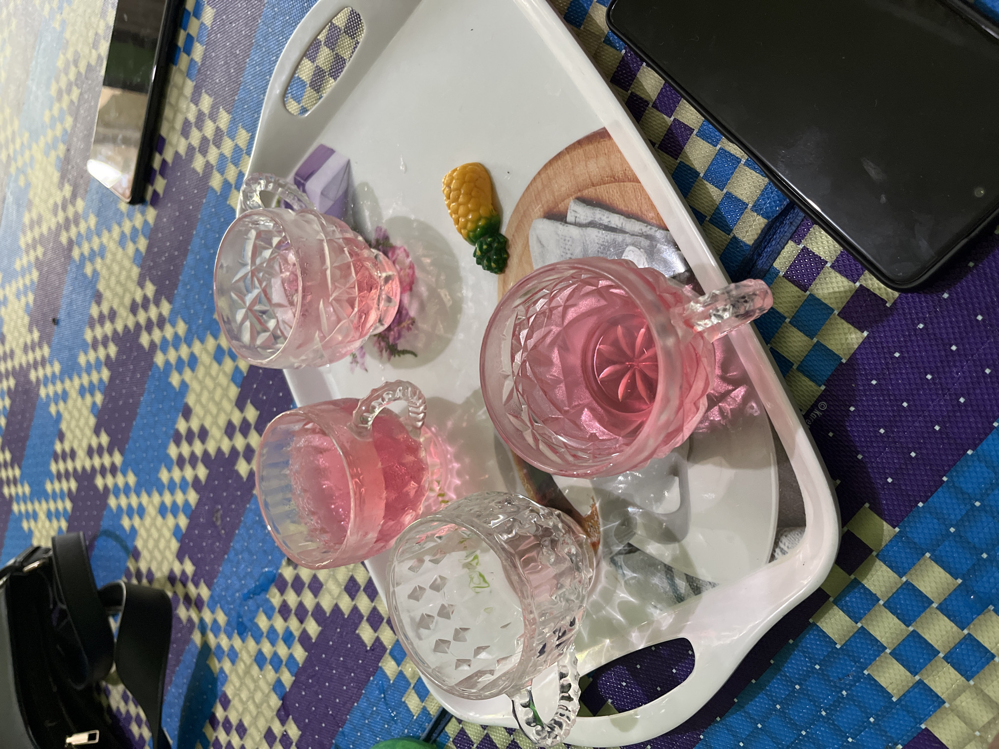 A white tray holding four ornate glass cups, three containing pink liquid and one empty, sits on a colorful woven mat. A small yellow and green pineapple-shaped candy rests on the tray next to a circular graphic of a napkin. A black handbag with a silver zipper and black straps is visible in the upper left corner. Two smartphones are present: one is partially visible in the upper right, and another is prominently displayed in the lower right. The scene is set in Babahjurong, Indonesia.