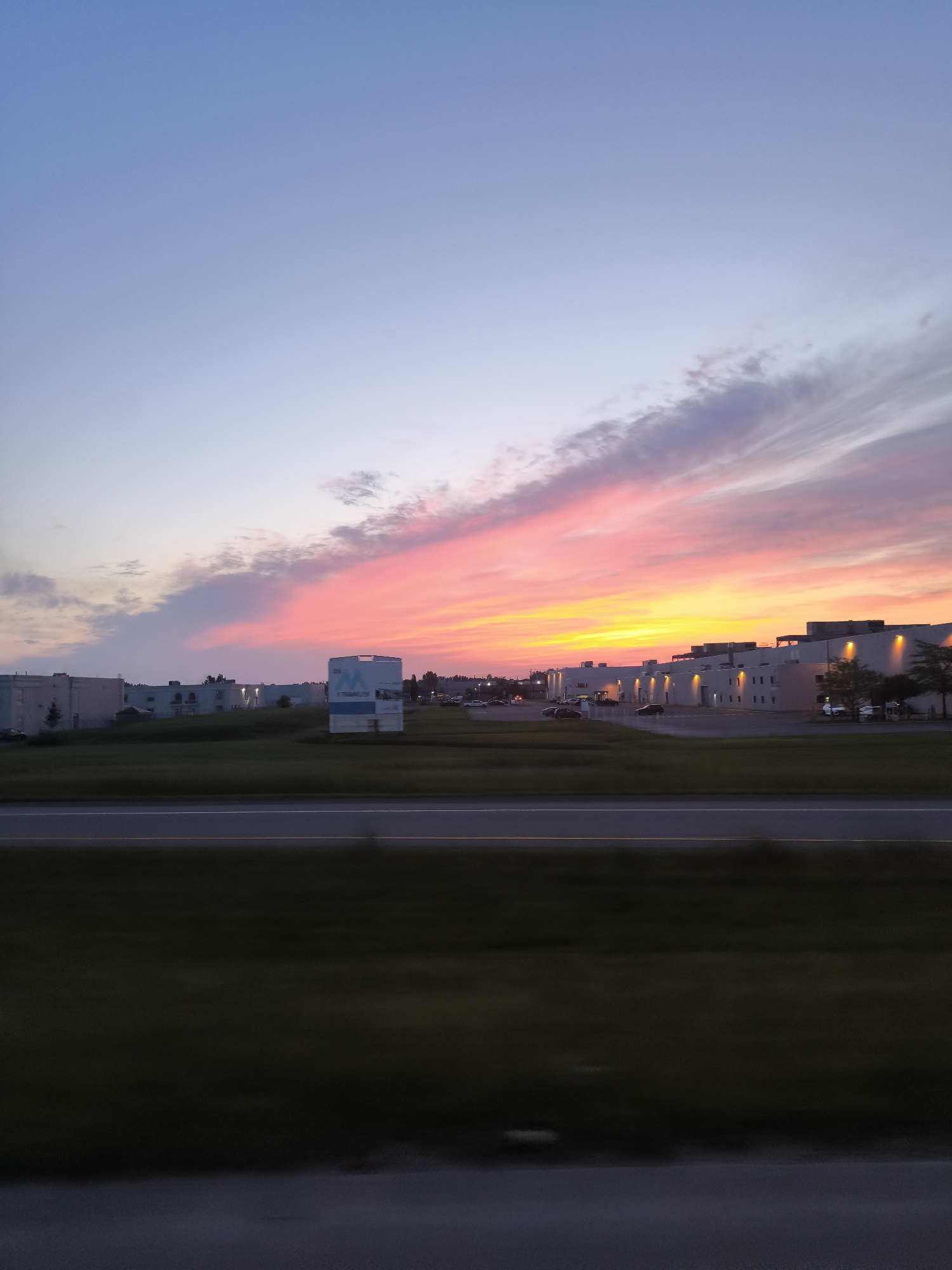 An exterior, full shot captures a sunset over a commercial area in Mirabel, Canada. The sky transitions from a soft blue at the top to vibrant hues of pink, orange, and yellow near the horizon, with wispy clouds scattered throughout. In the mid-ground, several low-rise industrial or commercial buildings with flat roofs line the landscape. One prominent building on the right features numerous evenly spaced lights illuminating its exterior walls. To the left of this building, a large white sign with a blue "M" logo is visible. A strip of green grass separates the buildings from a highway in the foreground, with a blurred motion effect suggesting the photo was taken from a moving vehicle. The highway itself is dark asphalt with white lane markings. The overall scene depicts a serene evening in an urban industrial setting.