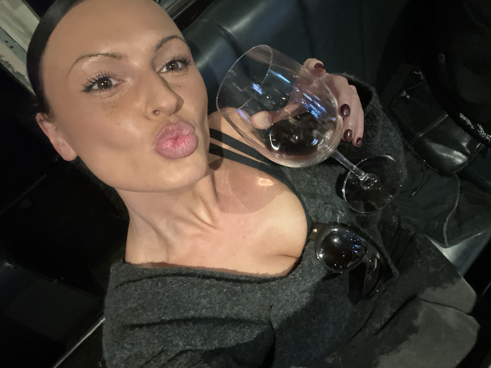 This close-up selfie features a woman from the chest up, striking a playful 'kissy face' pose with puckered lips. Her dark hair is neatly pulled back, drawing attention to her face, which has light brown eyes enhanced by dark mascara and eyeliner, and a scattering of freckles across her nose and cheeks. She is wearing a dark, possibly charcoal grey, fuzzy or knitted sweater or cardigan, beneath which a black top with visible straps can be seen.
In her left hand, she holds a wine glass filled with red wine. Her fingernails are painted a dark red or burgundy color. A pair of dark sunglasses with rounded frames is tucked into her clothing on her right side.
The background is dark and indistinct, suggesting an indoor, dimly lit setting such as a bar, lounge, or restaurant. Hints of dark, possibly upholstered furniture are visible behind her. The dark ambiance implies the photo was taken in the evening or at night. The activity depicted is a casual selfie, with the woman enjoying a drink. There is no visible text in the image. The setting aligns with an indoor venue in London, United Kingdom.