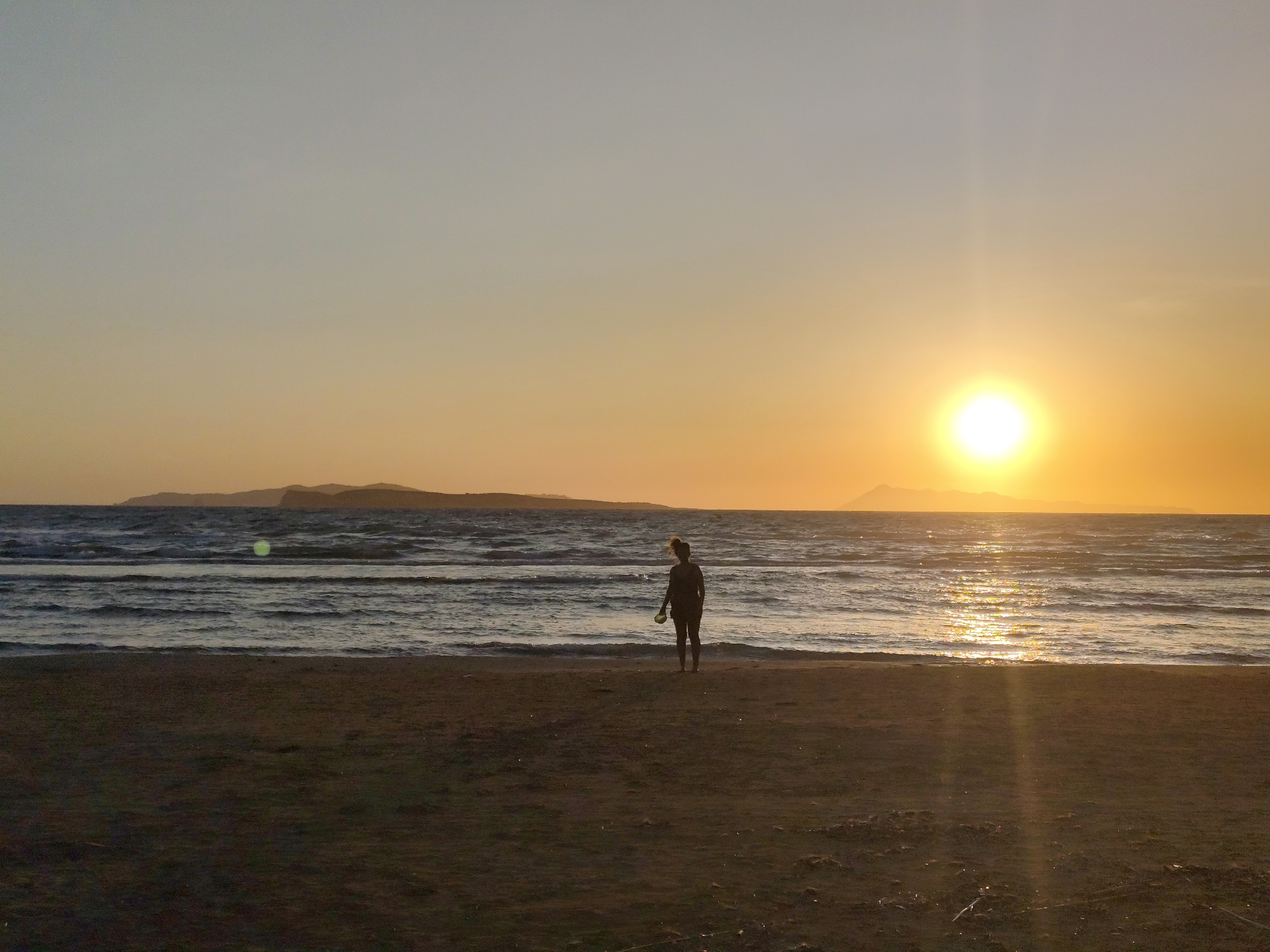 A woman stands on a sandy beach in Agios Stefanos, Greece, facing the ocean. The sun is setting, casting a golden glow over the water and sky. In the distance, a silhouette of mountains is visible. The sea is choppy with small waves.
