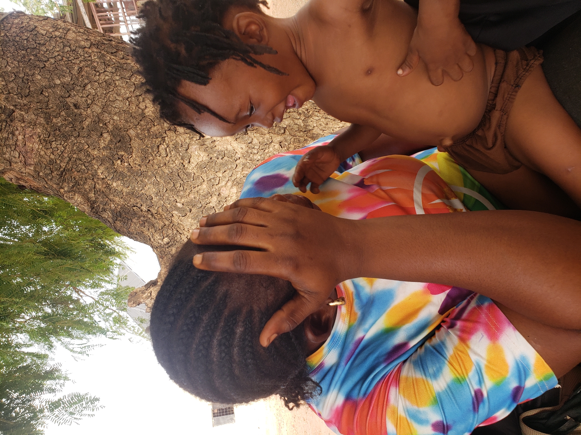 This media file captures an intimate moment between an adult and a young child in Adankolo, Nigeria. The scene is set outdoors, likely under a large tree, as evidenced by the prominent textured tree trunk on the left side of the frame and green foliage visible in the background against a bright sky.
The adult, with dark skin and braided hair, is positioned on the right, wearing a colorful tie-dye top featuring shades of blue, pink, orange, and yellow. Their right hand is placed over their head or forehead, suggesting a moment of rest, contemplation, or perhaps a playful gesture in response to the child. A gold earring is visible in their right ear.
The child, also with dark skin and dreadlocks, is shirtless and wears brown bottoms, leaning over the adult. Their expression is lively and joyful, with their mouth open as if mid-laughter or speaking. One of the child's hands rests near the adult's waist. The interaction between the two appears affectionate and playful.
The lighting indicates bright daylight, suggesting a warm day outdoors. The ground around the tree trunk appears dry and cracked in some areas. There is no visible text in the image.