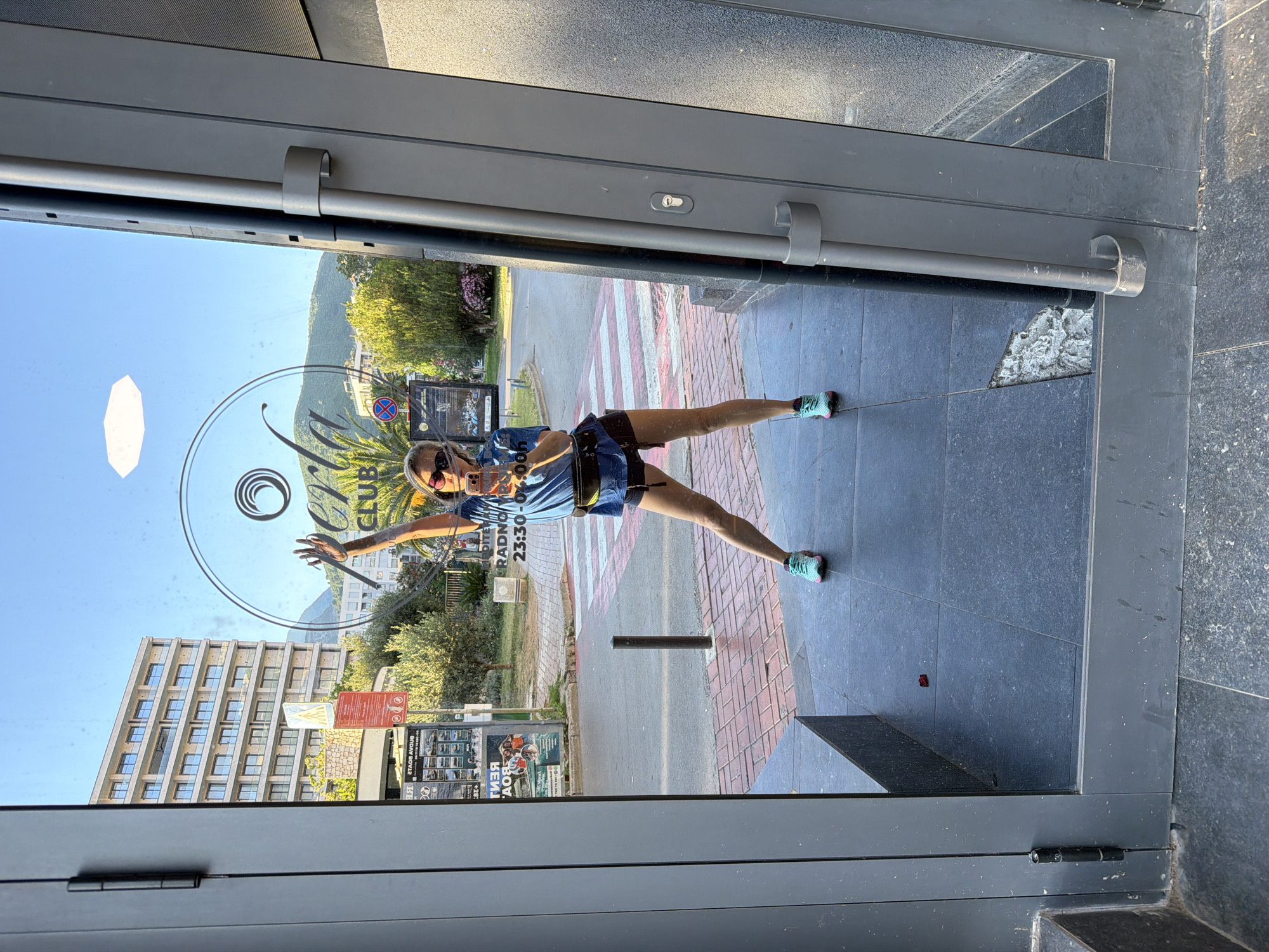 A woman is taking a mirror selfie in front of a glass door. She is wearing a blue t-shirt, black shorts, and turquoise sneakers. She is holding up her left hand and smiling at the camera. The glass door has a logo that says "Perla Club" and the hours "23:30-00:00". In the background, there is a building, trees, and a street with a crosswalk. The scene appears to be in Budva, Montenegro, indicated by the "Budva Boats" advertisement visible in the reflection. The woman is standing on a tiled floor.