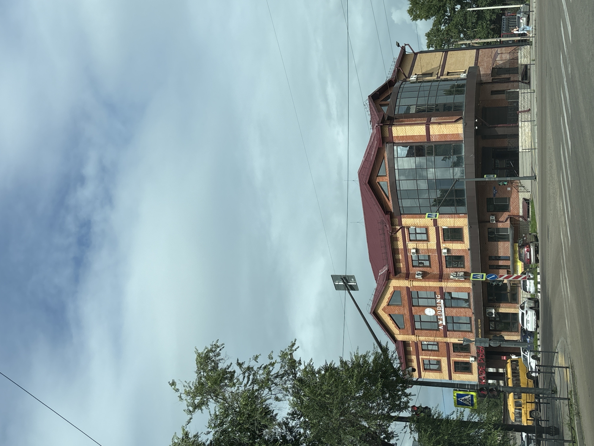 An exterior, eye-level, medium shot captures a three-story brick building with a red roof on a cloudy day in Blagoveshchensk, Russia. The building, labeled "A.WEDDING" in white lettering, has a rounded glass facade on the first and second floors. Several cars are parked on the street in front of the building. A yellow school bus is parked to the left of the frame. A pedestrian crossing signal is visible on the left side of the image. Trees line the street, and power lines are strung across the sky. The overall impression is that of a street scene in a Russian city.