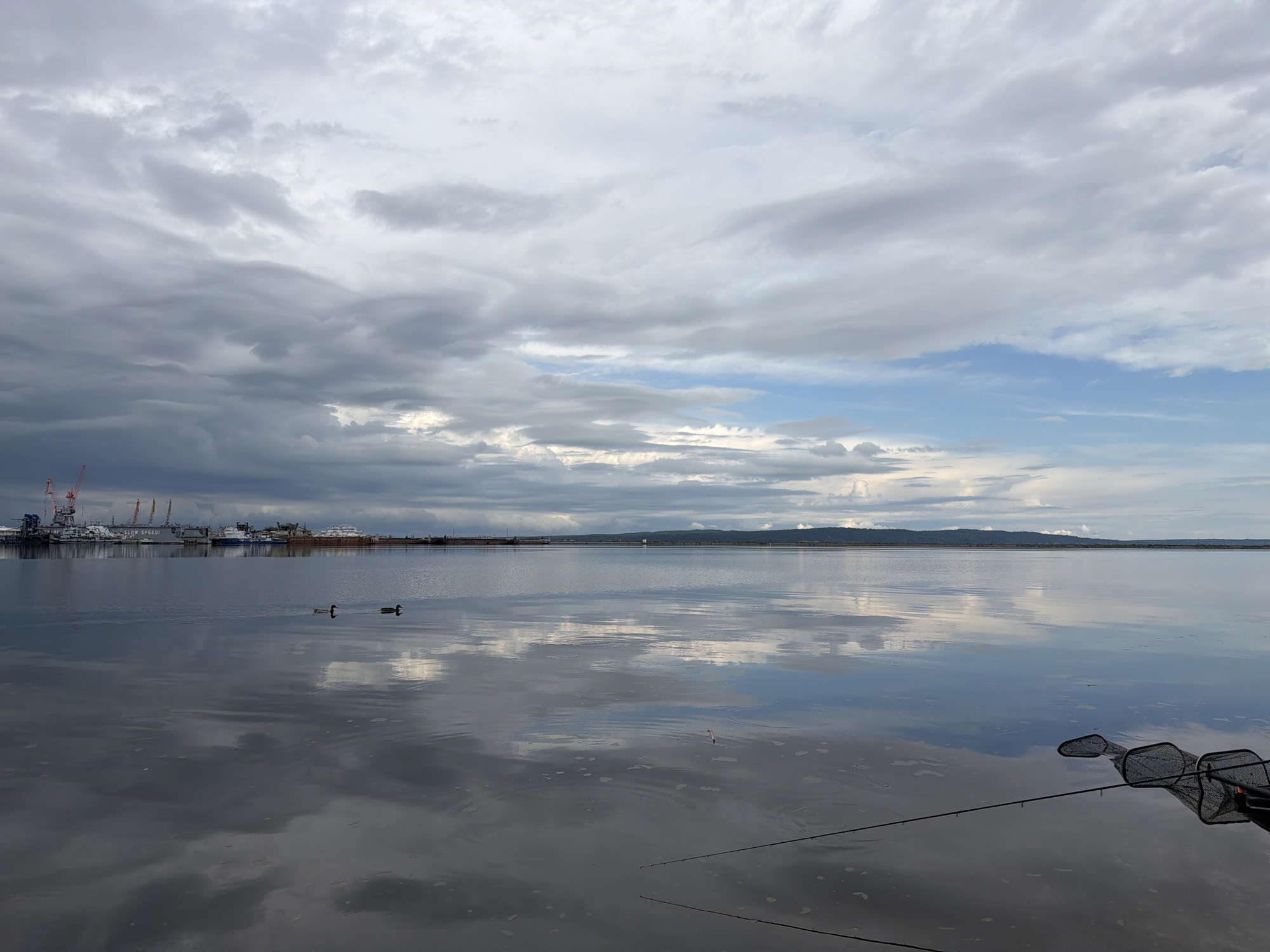 The image shows a calm body of water reflecting a cloudy sky. In the distance, there are port cranes and various boats docked at a harbor. Two ducks are visible swimming in the water. Fishing nets and fishing poles are in the foreground. The location is the city of Chaikovsky, Russia.