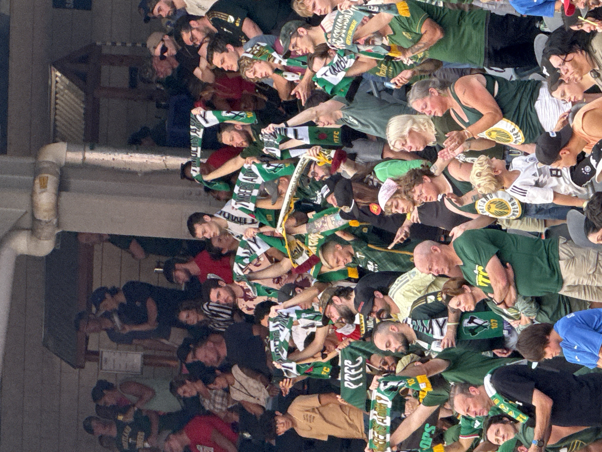 The image depicts a crowded outdoor stadium, likely during a sporting event, with spectators of various ages and ethnicities visible. The setting appears to be a sunny day, with the crowd shaded by an overhang. Many individuals are wearing green and white clothing, some adorned with team scarves and banners, suggesting a shared affiliation with a sports team. The atmosphere appears enthusiastic, with some individuals holding up scarves and banners, possibly in support of their team. The crowd is densely packed, indicating a popular event. The background shows stadium architecture, including seating sections and structural elements. The individuals are casually dressed, appropriate for an outdoor event.