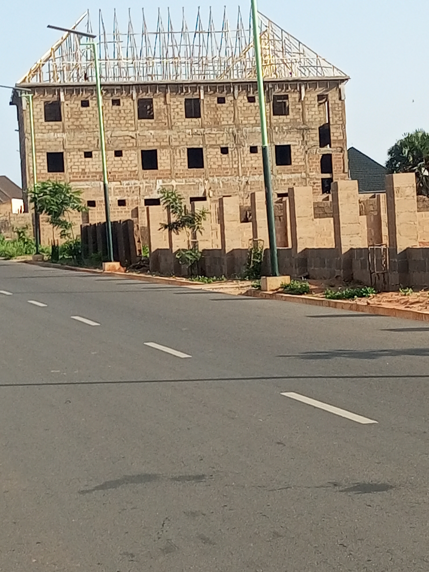 The image shows a multi-story building under construction on a street in Garko, Nigeria. The building is made of brick and appears to be several floors high, with exposed rebar on the roof, indicating it is not yet completed. A dark-colored roof is visible on a lower structure adjacent to the main building. The street in the foreground is paved asphalt with white lane markings. Several streetlights are present, with one prominently positioned in front of the construction. Trees and vegetation are scattered around the building site, and some construction materials, possibly bricks or concrete blocks, are visible along the perimeter of the property. The sky is clear and blue, suggesting fair weather. There are no people or vehicles visible in the image. The scene is an urban environment with ongoing construction work.