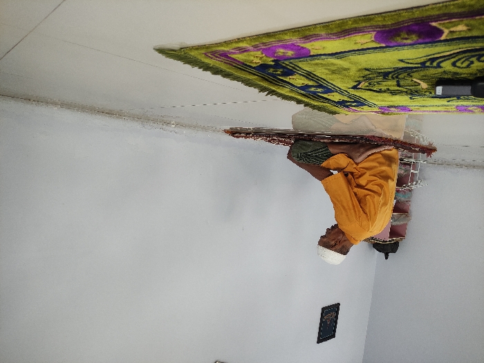 An indoor scene captures an adult man engaged in Islamic prayer, kneeling on a prayer mat in a simple room. The man, with grey or white facial hair, wears a white skullcap (kufi) and an orange-yellow long-sleeved shirt over green or patterned lower garments, possibly a sarong. He is positioned facing right in the image, with his body in a focused, reverent posture typical of *salat*, likely *jalsa* (sitting between prostrations) or a similar kneeling position.
He is situated on a small, reddish-brown patterned prayer mat, which is placed atop a larger, vibrant green prayer mat featuring intricate purple and dark blue floral or geometric patterns. To his right, on the green mat, a small dark rectangular object (possibly an electronic device or speaker) is visible. Behind him, a white and green striped woven bag or basket is partially obscured.
The setting is a plain room with white walls, creating a minimalist backdrop for the spiritual activity. A corner where two walls meet is clearly visible, and a small, dark-framed object, possibly a picture or religious scripture, hangs on the wall in the lower left of the frame. The lighting appears soft and ambient, suggesting an indoor environment during an unspecified time of day. The scene evokes a sense of peace and devotion, consistent with prayer in a private or semi-private setting in Situbondo, Indonesia. No discernible text is visible on the framed object due to distance and resolution.