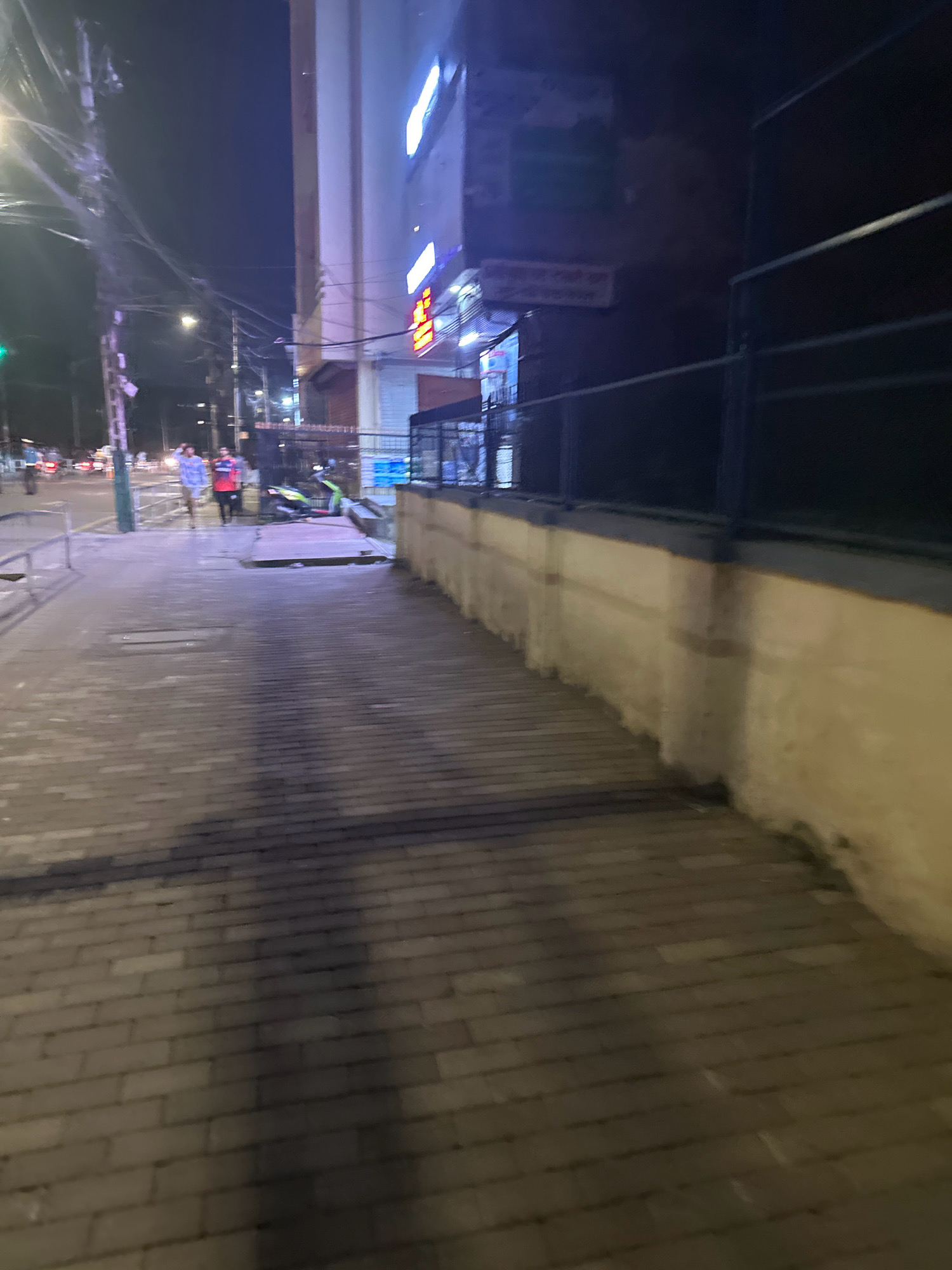The image depicts a street scene at night in Kathmandu Metropolitan City, Nepal. A paved sidewalk runs diagonally from the lower left to the upper right corner of the frame. The sidewalk is made of rectangular brick-like pavers, with a shadow cast across it from an unseen light source. To the right of the sidewalk is a low, light-colored concrete wall topped with a dark metal fence. Beyond the wall, the facade of a building is visible, illuminated by bright neon signs and internal lighting, suggesting commercial establishments. The building's architecture appears modern, with multiple levels. In the mid-ground, on the street bordering the sidewalk, two individuals are walking. One, on the left, is wearing a checkered shirt and shorts, while the other, on the right, is in a red and black outfit. A green motorcycle is parked on the street near them. In the background, the street continues with traffic lights and more street lighting, indicating an urban environment with ongoing activity despite the late hour. The overall scene is dimly lit by artificial sources, characteristic of a city at night.