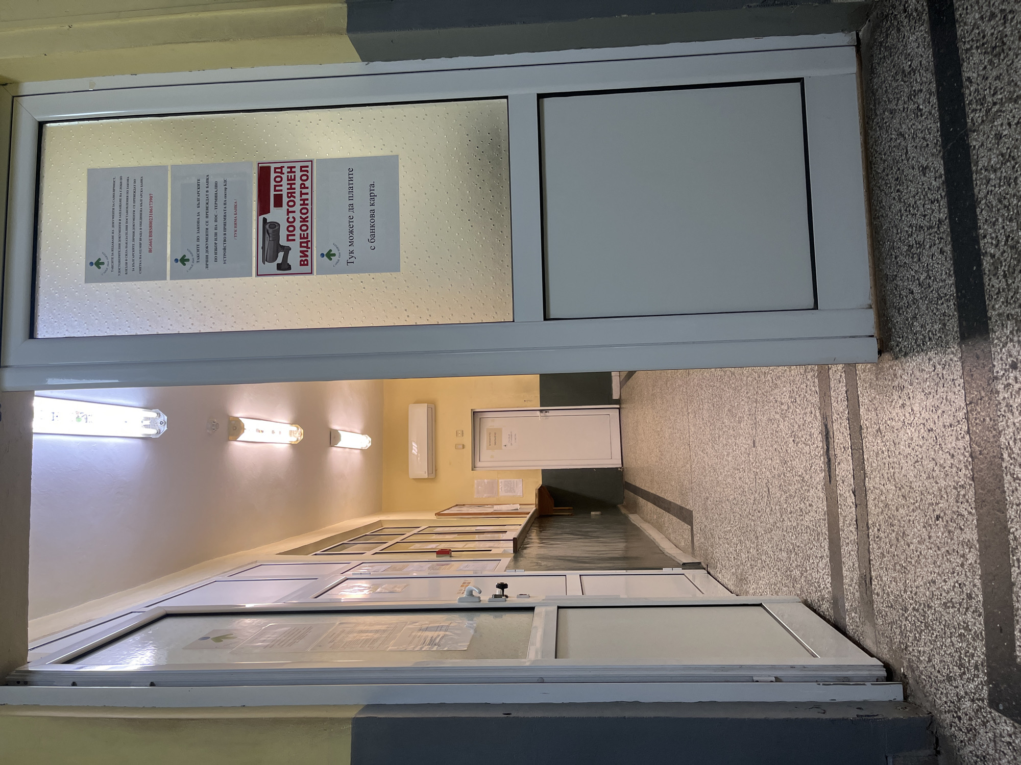 The image shows a hallway with a door on the right side of the frame. On the door, there are several signs. One sign indicates "ПОД ПОСТОЯНЕН ВИДЕОКОНТРОЛ," which translates to "UNDER CONSTANT VIDEO SURVEILLANCE." Another sign states "Тук можете да платите с банкова карта," meaning "Here you can pay by bank card." The hallway appears to be inside a building, possibly a government office or a bank, given the official-looking signs. The lighting in the hallway is provided by fluorescent lights on the ceiling. The walls are painted yellow and gray. The floor is made of speckled terrazzo. The open door on the left suggests an entrance to another area or room. The scene does not contain any identifiable people or specific landmarks of Vratsa, Bulgaria.